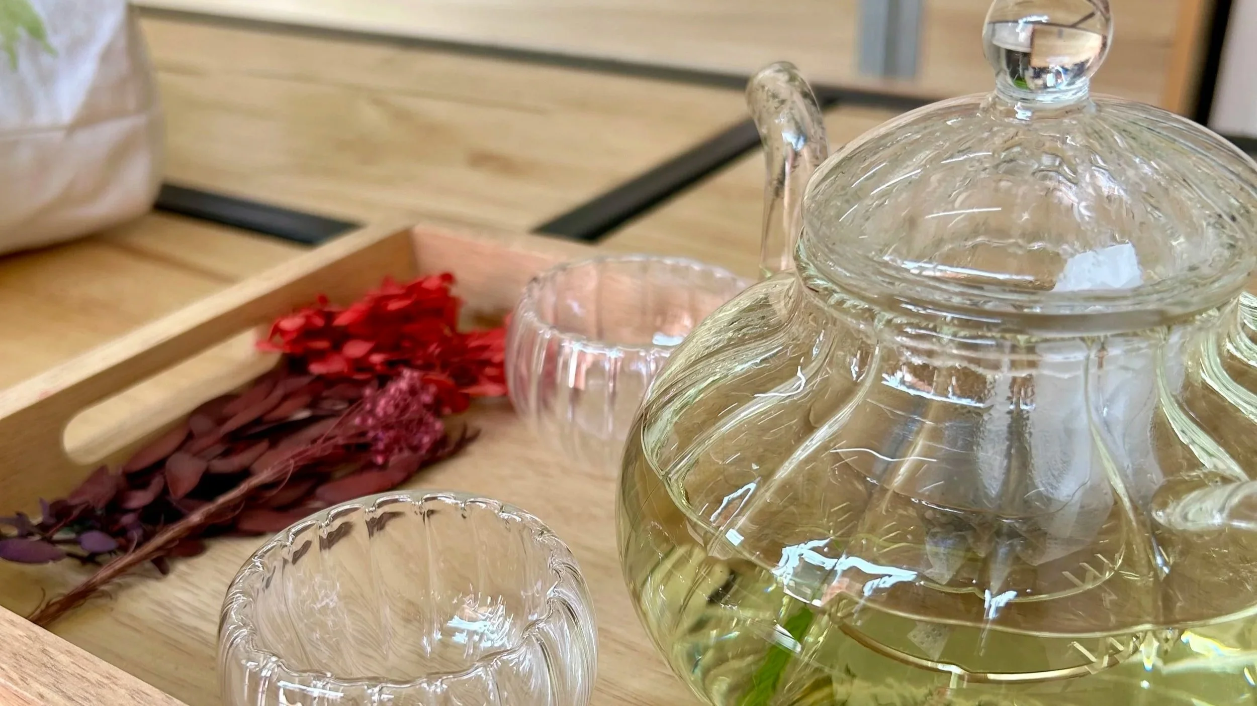 A glass teapot filled with tea, two small glass teacups, and a tray with dried red and purple flowers on a wooden table.