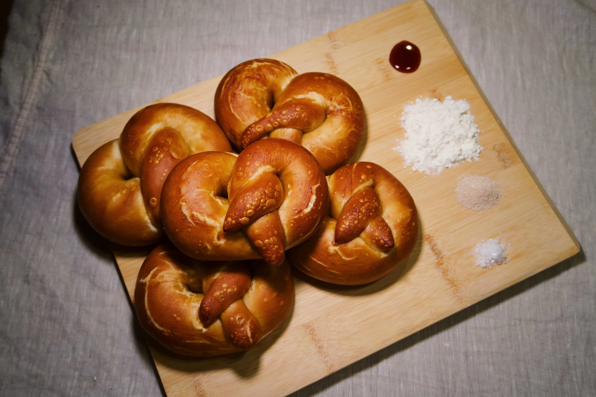 Six soft pretzels on a wooden cutting board with salt, flour, a small amount of cinnamon, dark syrup, and another powder on the side.