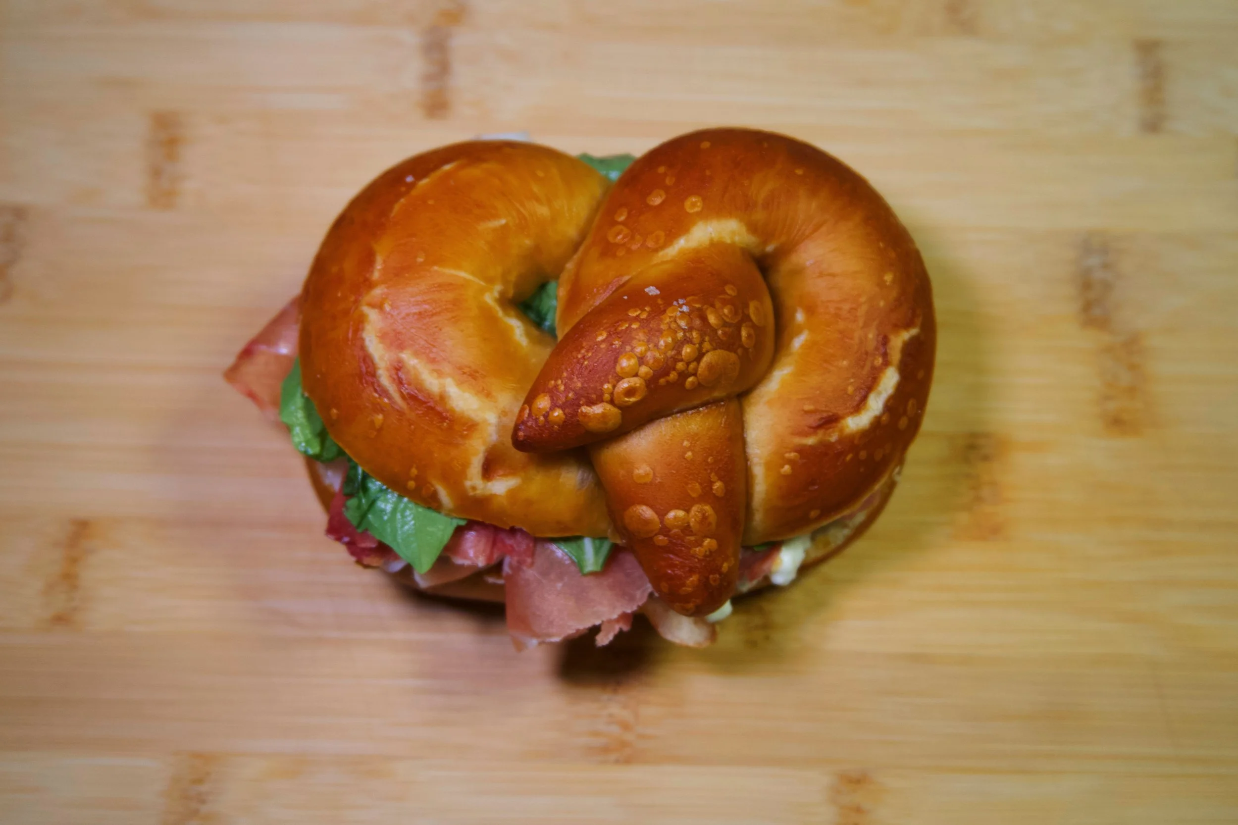 Baked pretzel roll sandwich filled with lettuce, sliced meats, and toppings on a wooden surface.