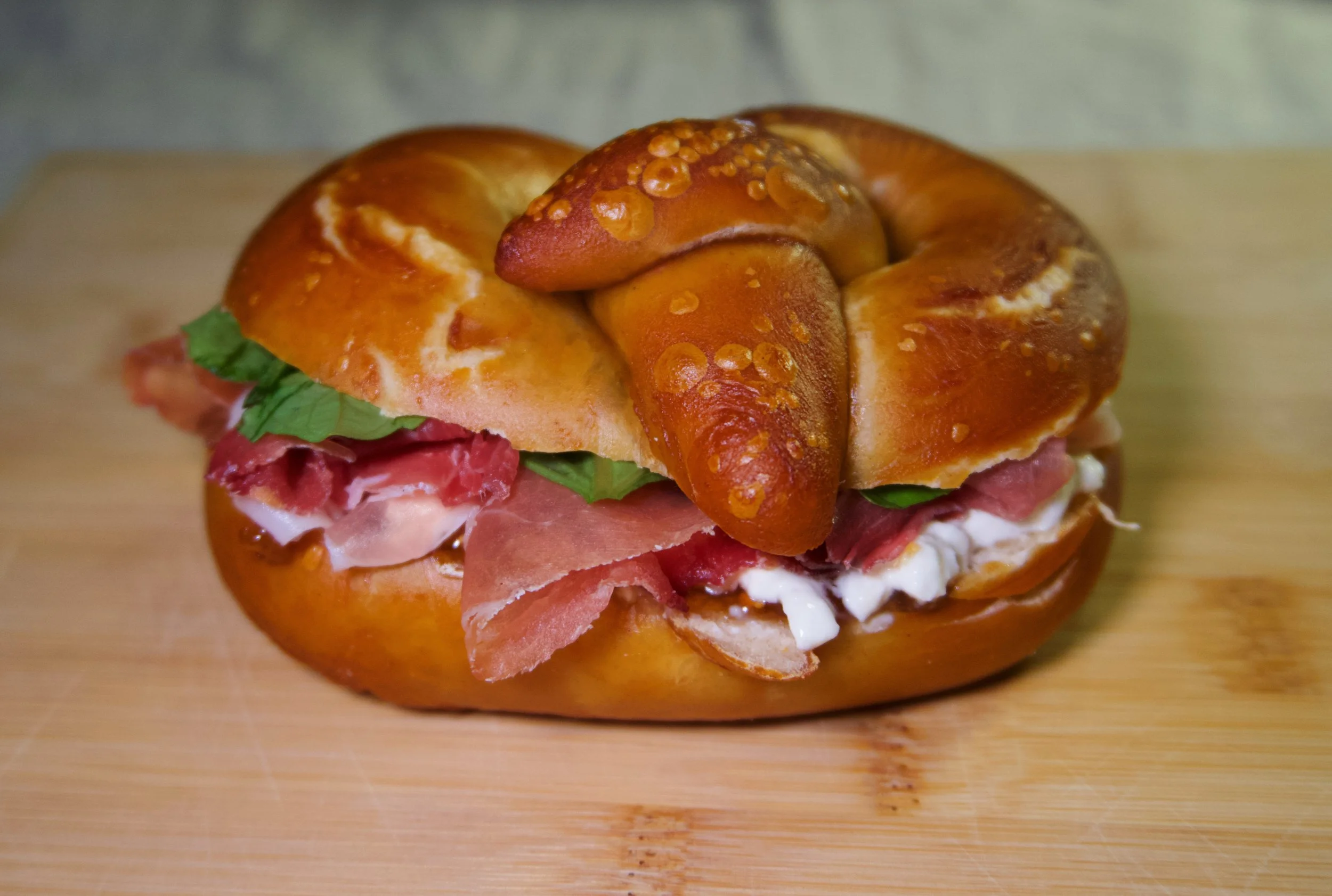 A sandwich on a wooden cutting board with prosciutto, lettuce, and cheese inside a pretzel bun
