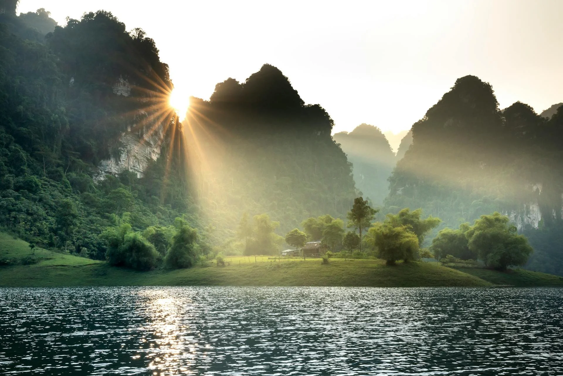Sun at sunrise shining through mountains and trees over a river and grassy landscape.