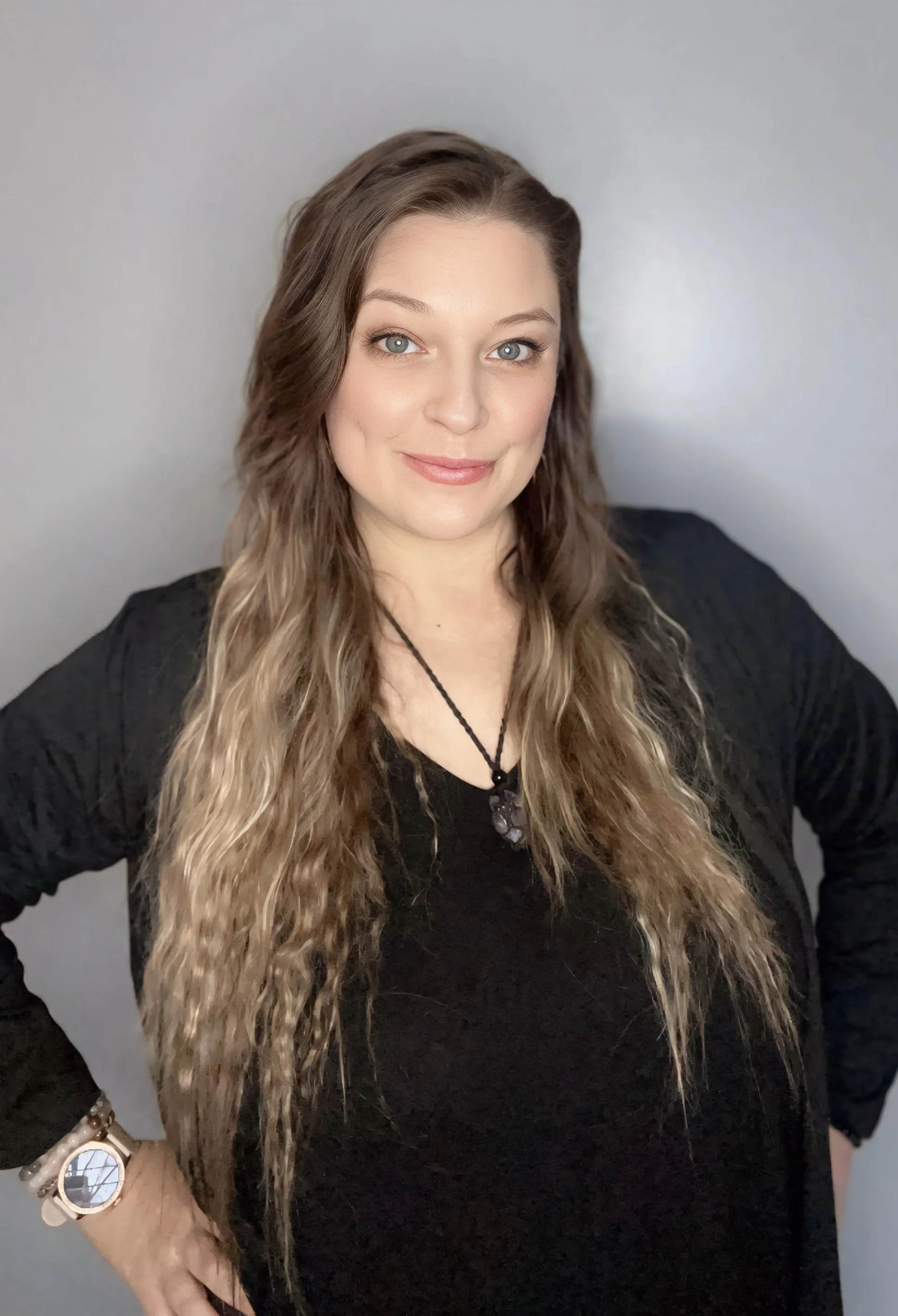 A woman with long, wavy blonde hair, light skin, and blue eyes, wearing a black top, a black necklace, and a watch with a white strap, is smiling and posing in front of a plain gray background.
