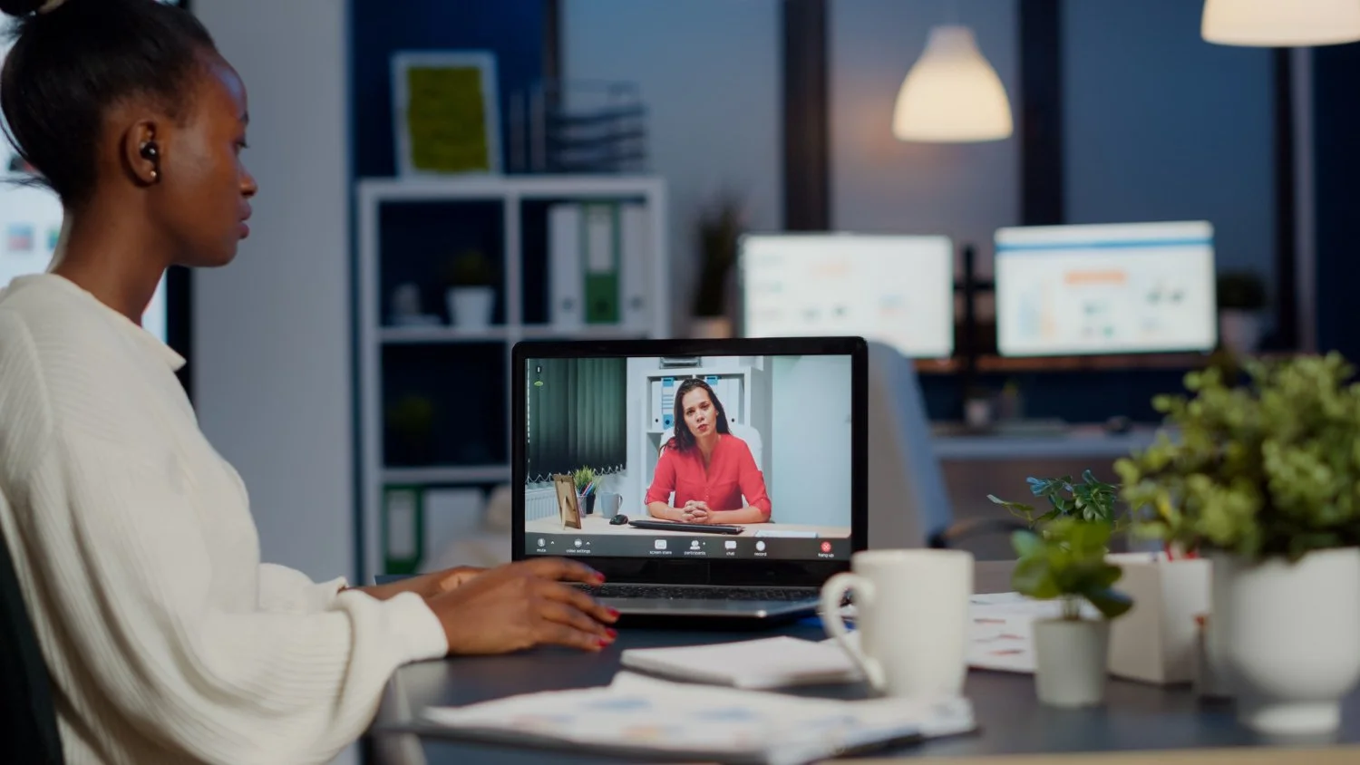 A woman participating in a video conference at her office desk, with a computer screen showing another woman in a red sweater.