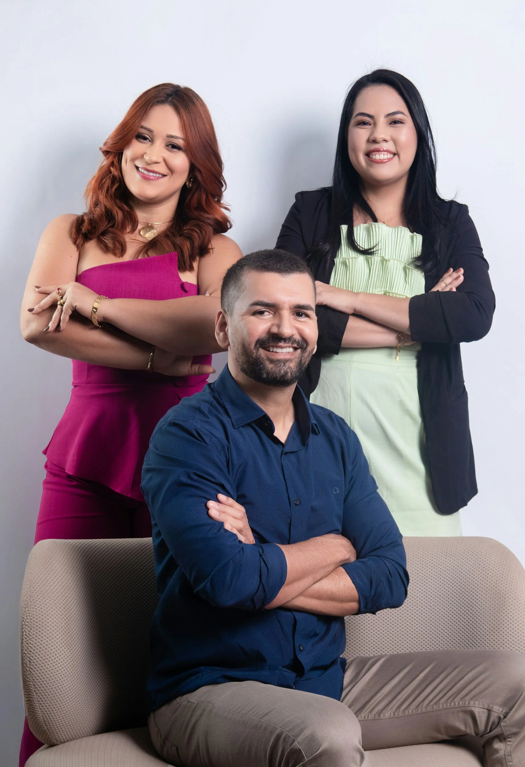 Three smiling adults, two women and one man, posing for a photo against a plain white background. The man is sitting on a beige couch with his arms crossed, wearing a blue shirt. One woman with dark hair is standing behind him with her arms crossed, wearing a black blazer and a light green dress. The other woman with red hair is standing next to him with her arms crossed, wearing a bright pink dress.