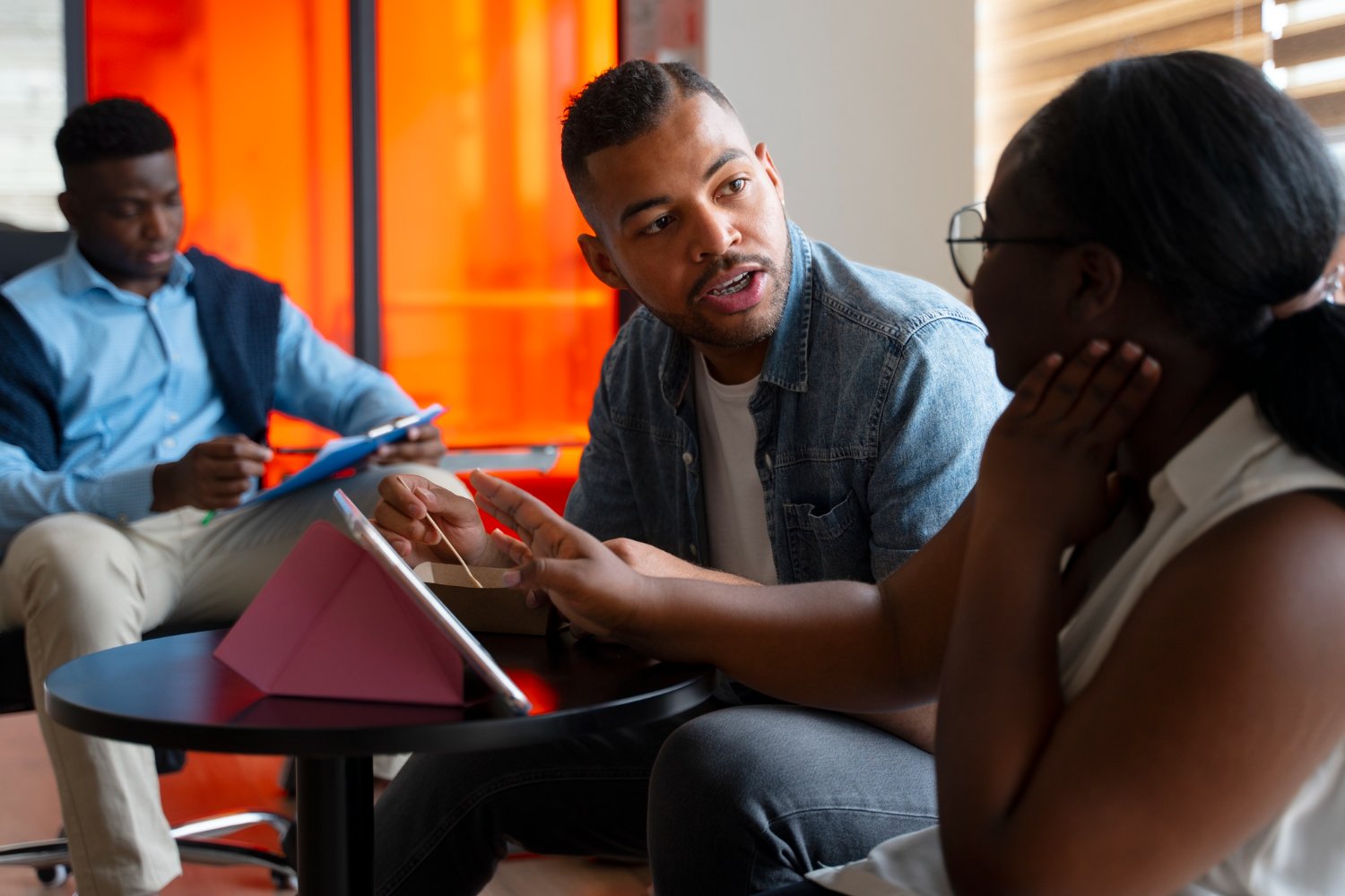 Three young adults, two men and one woman, having a conversation in a modern, brightly lit room with orange accents. The woman is listening attentively while the man in the denim jacket is speaking.