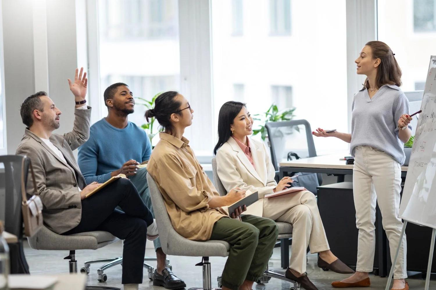 A woman presenting to a diverse group in a modern office setting, with four people seated and listening attentively, one raising his hand.