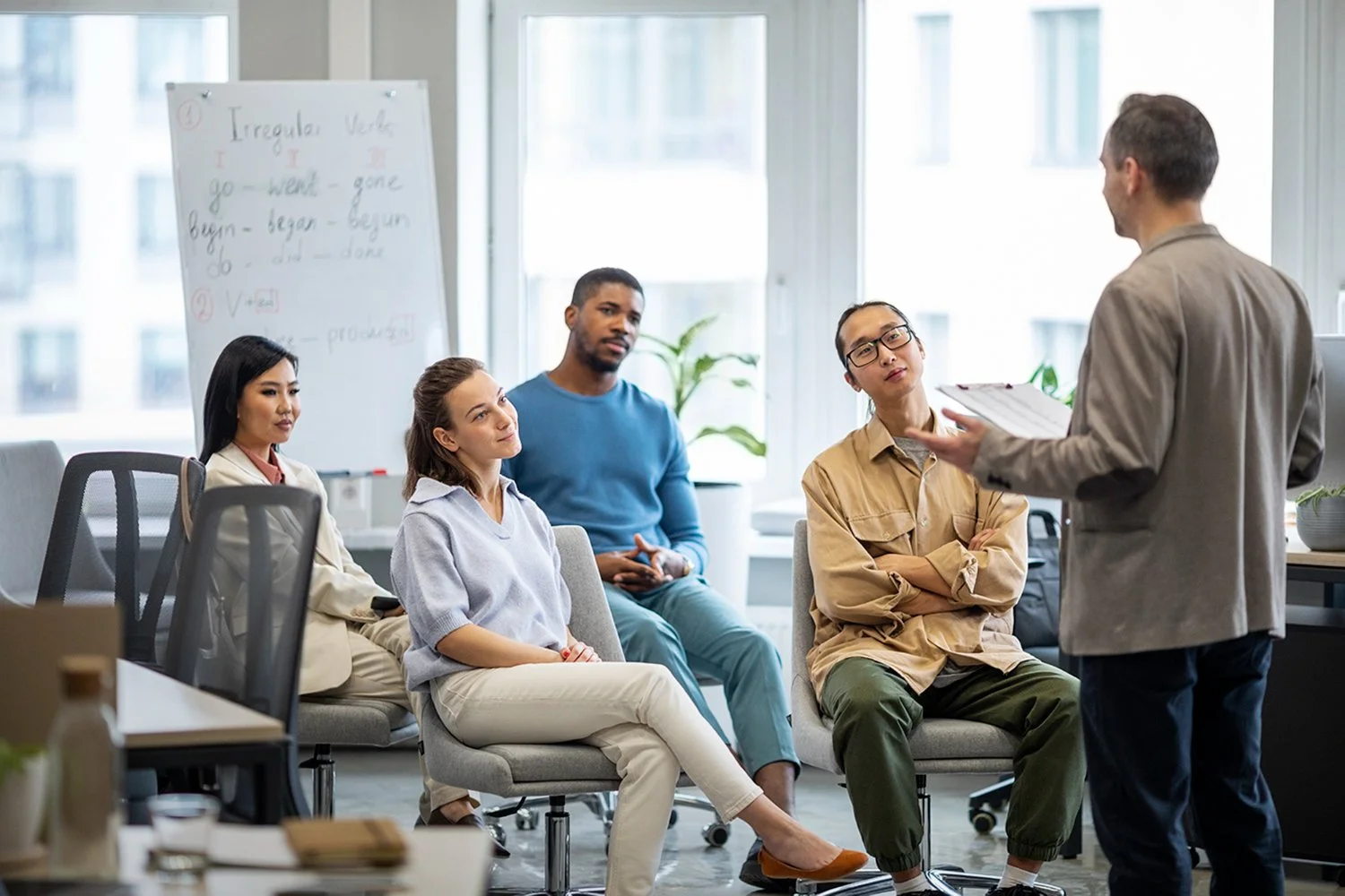 A diverse group of five people attending a meeting or class in a modern office. One person is standing and presenting while the others are seated and listening attentively, with a whiteboard and large windows in the background.