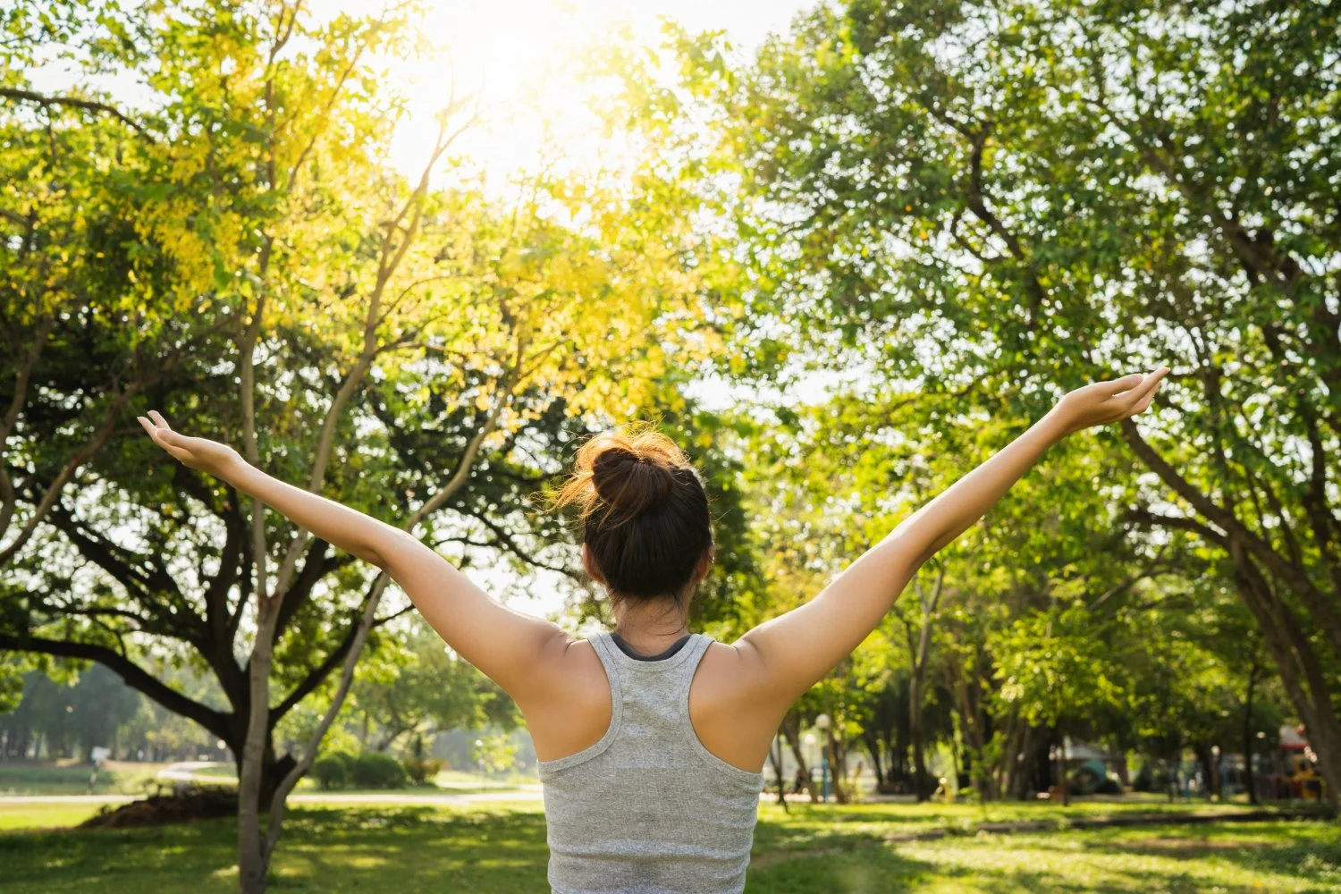 A woman stands in a park with her arms raised open towards the sunlight and trees in the background.