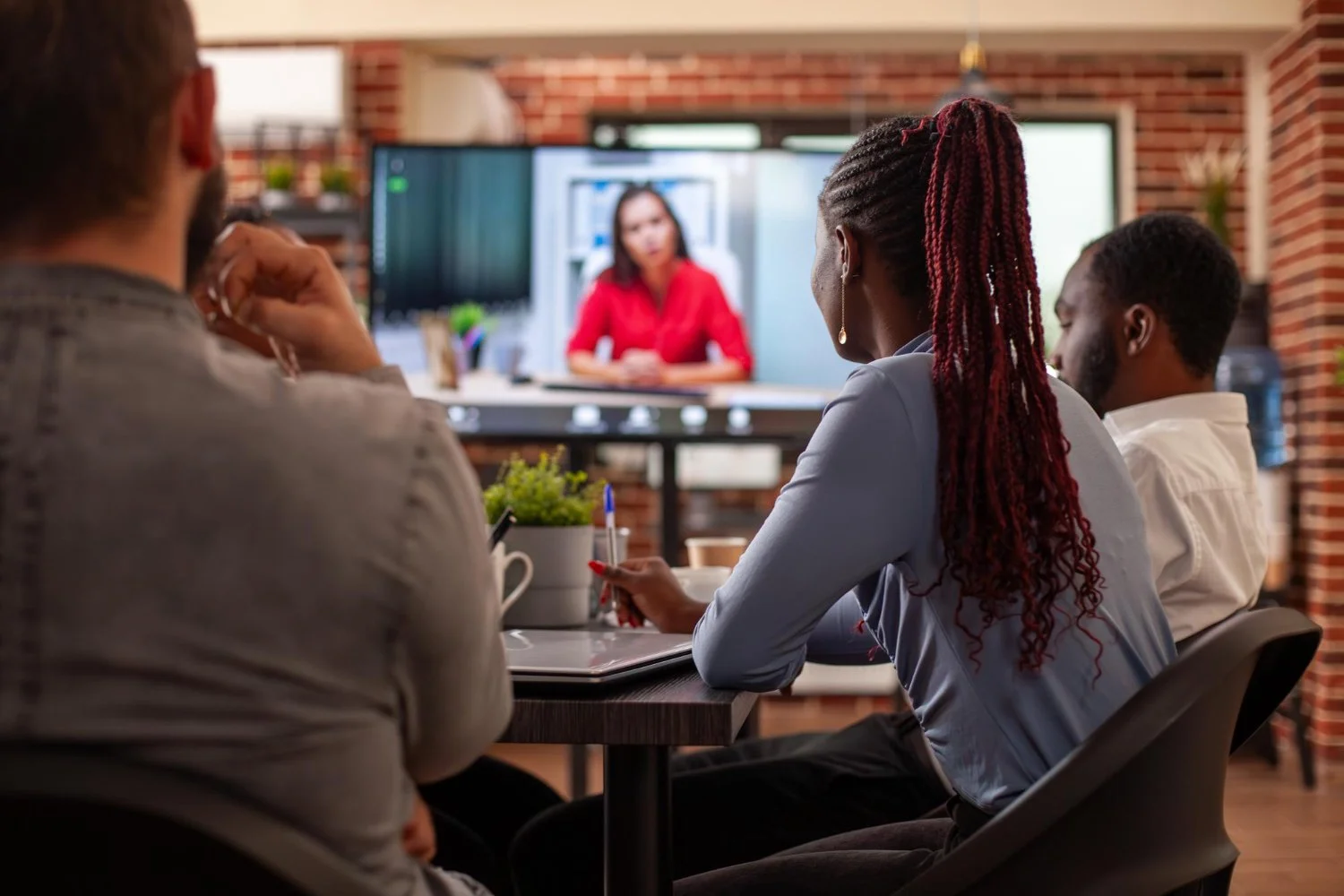People attending a virtual meeting in a conference room, viewing a woman speaking on a large screen.