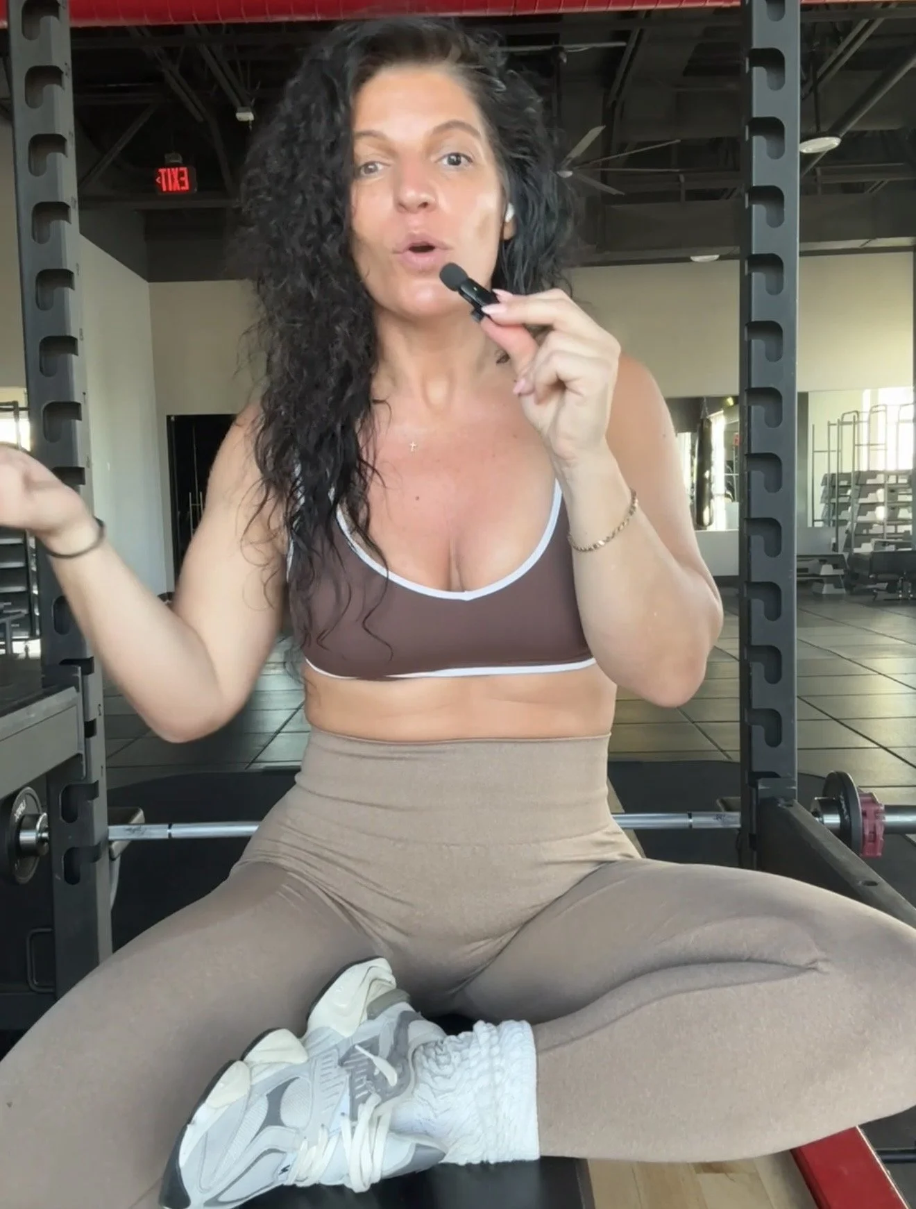 A woman with curly dark hair in workout attire speaking into a microphone in a gym. She is sitting with her legs crossed, wearing a brown sports bra, beige workout pants, and gray and white sneakers. The gym has weightlifting equipment and an exit sign visible in the background.