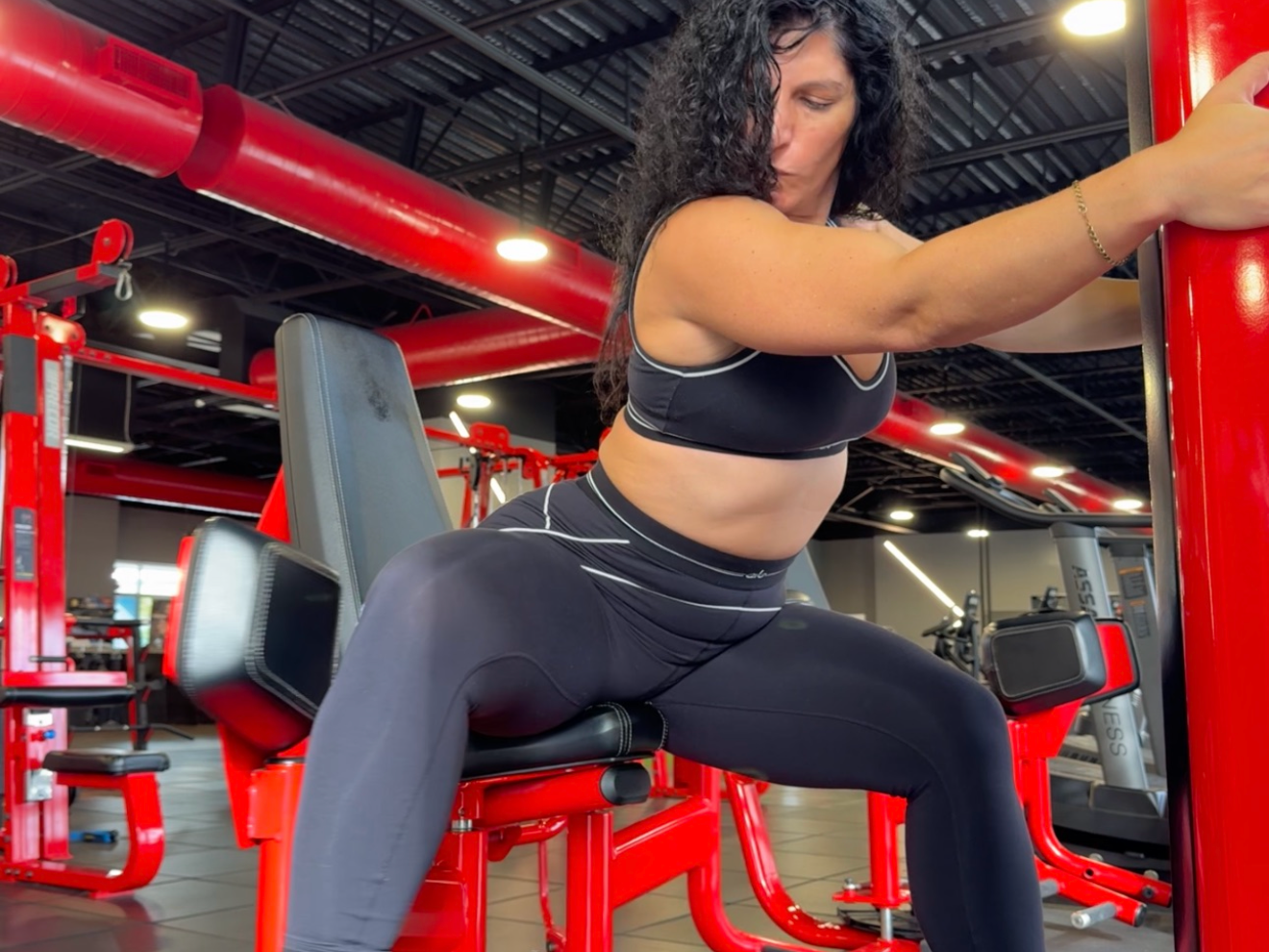Woman in black sports bra and leggings working out on a leg curl machine at a gym with red equipment and black ceiling.