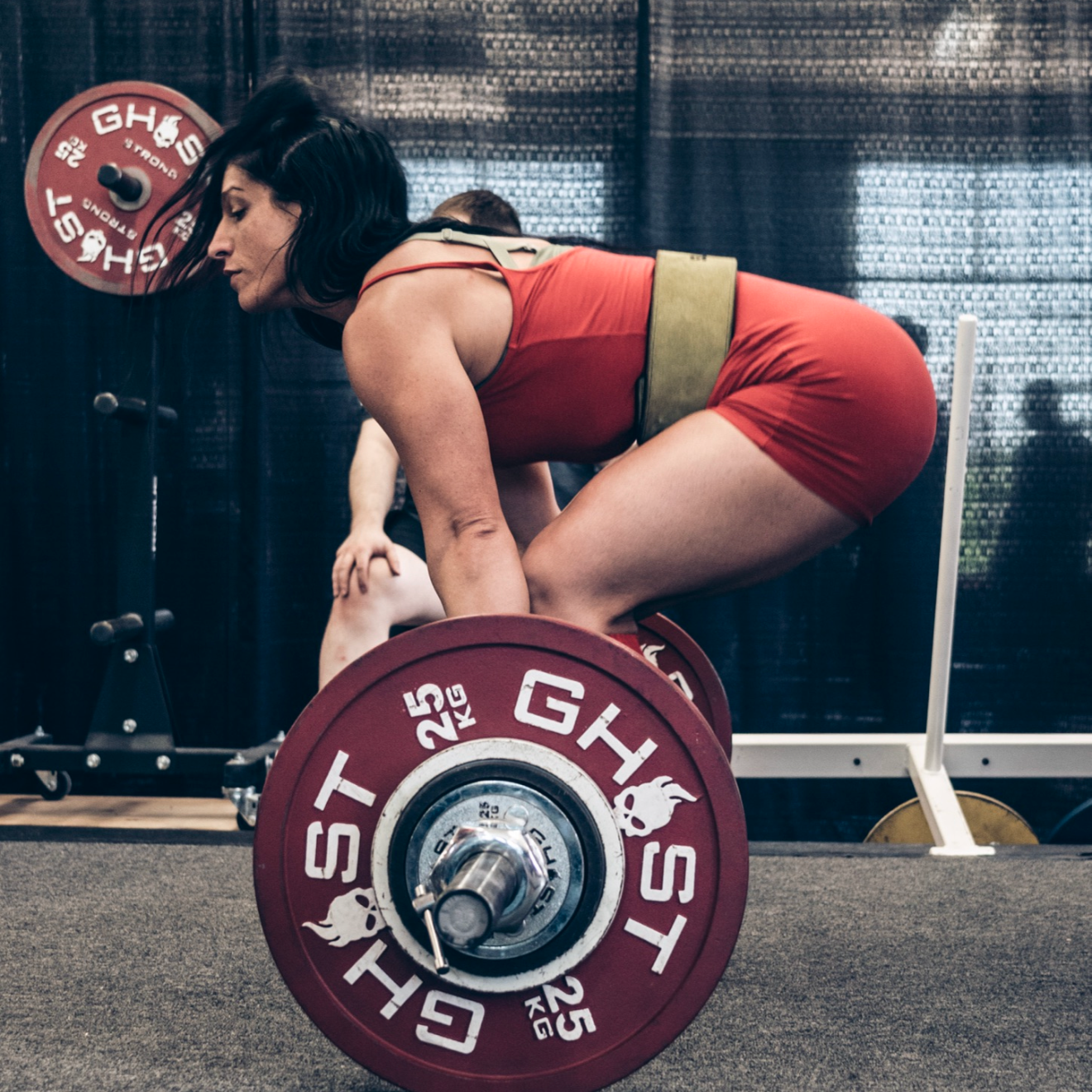 A woman in red sportswear performing a deadlift with a barbell loaded with red weight plates in a gym.