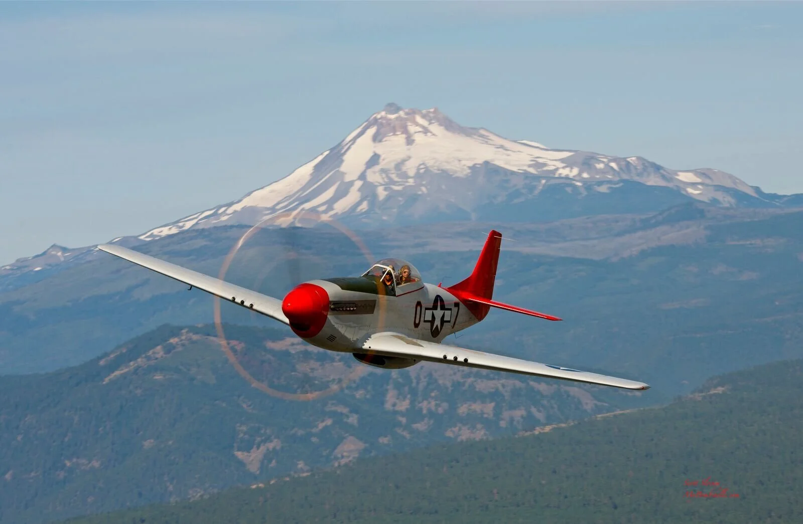 A vintage fighter plane flying in front of a snow-capped mountain with a rainbow halo around its propeller.
