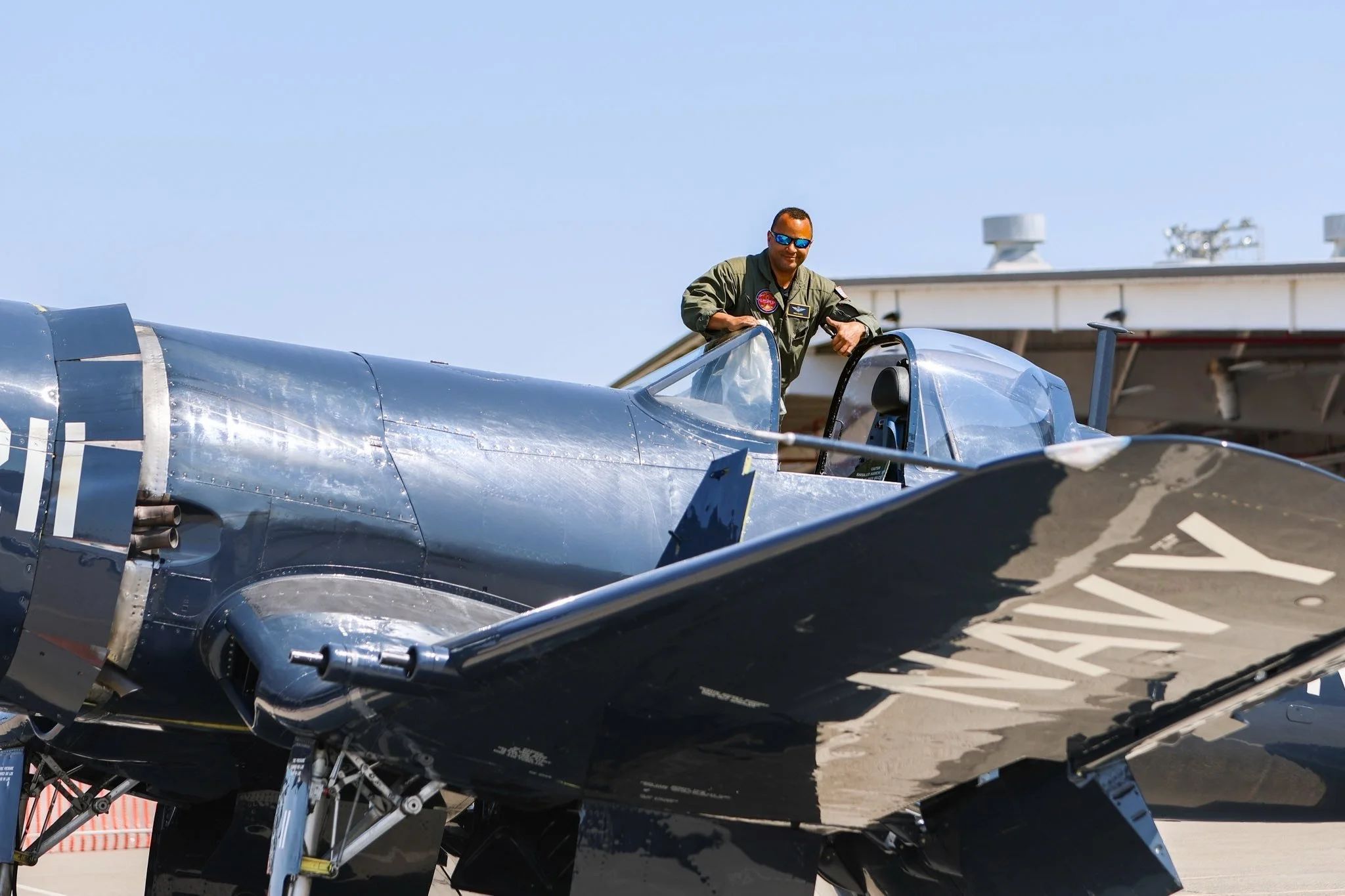A pilot standing next to a military jet aircraft on the tarmac, wearing sunglasses and a green flight suit.