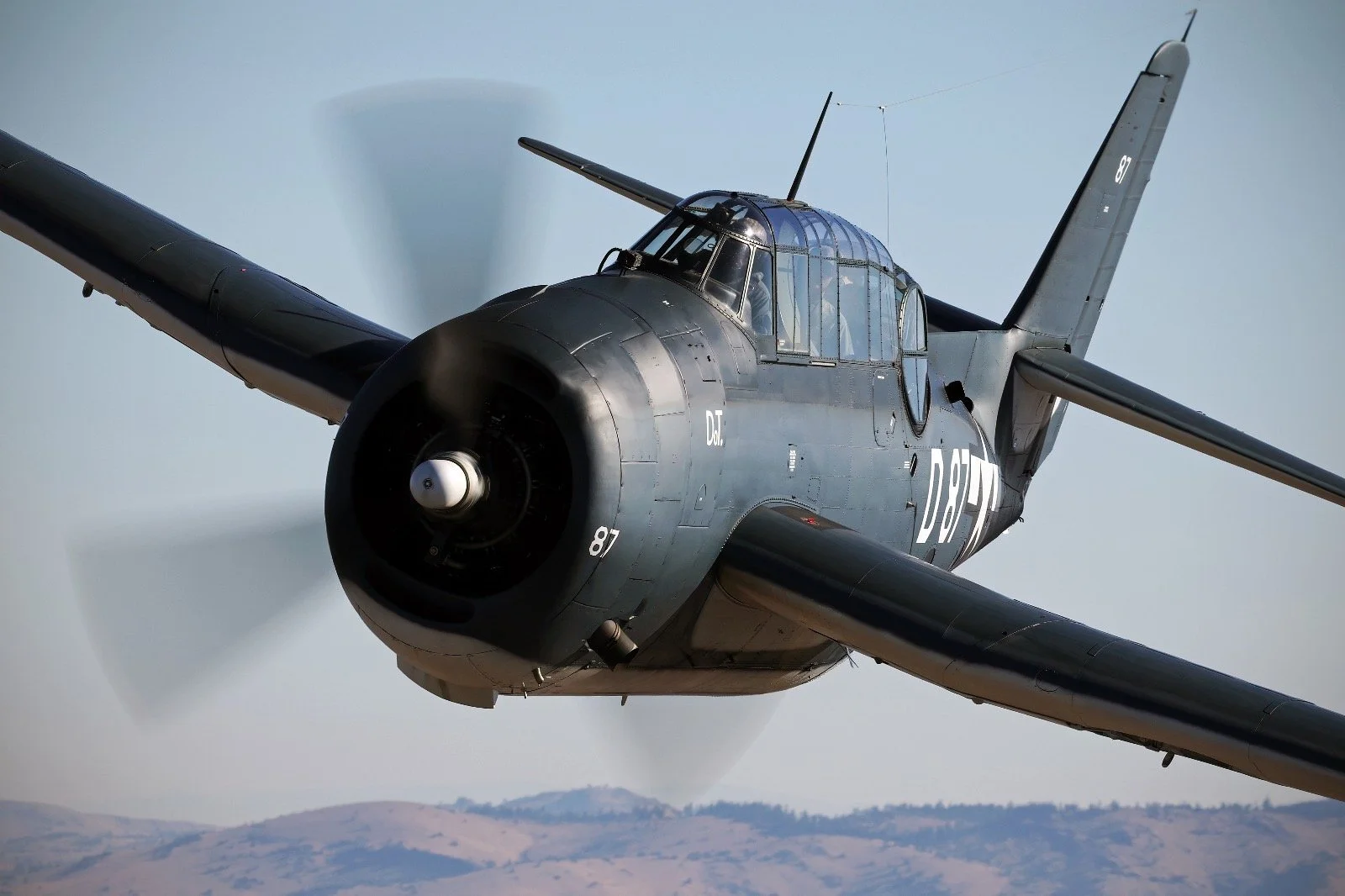 A vintage military aircraft flying through the sky with mountains visible in the background.