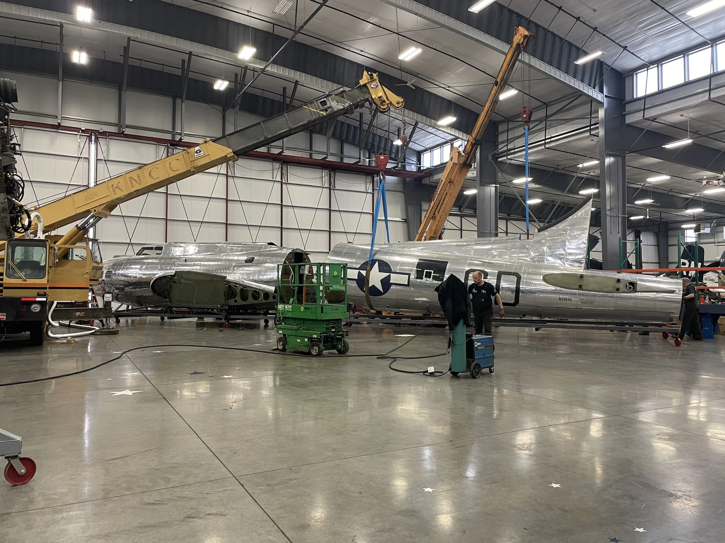 An indoor aircraft hangar with workers and equipment surrounding a vintage silver airplane with U.S. military markings.