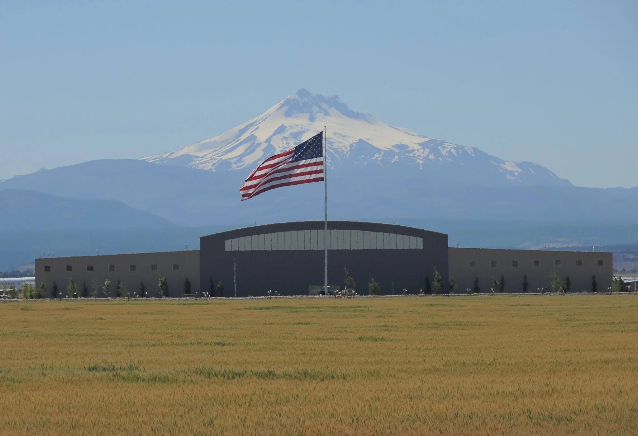 View of a large snow-capped mountain in the background with a building and a waving American flag in the foreground, and a grassy field in front.