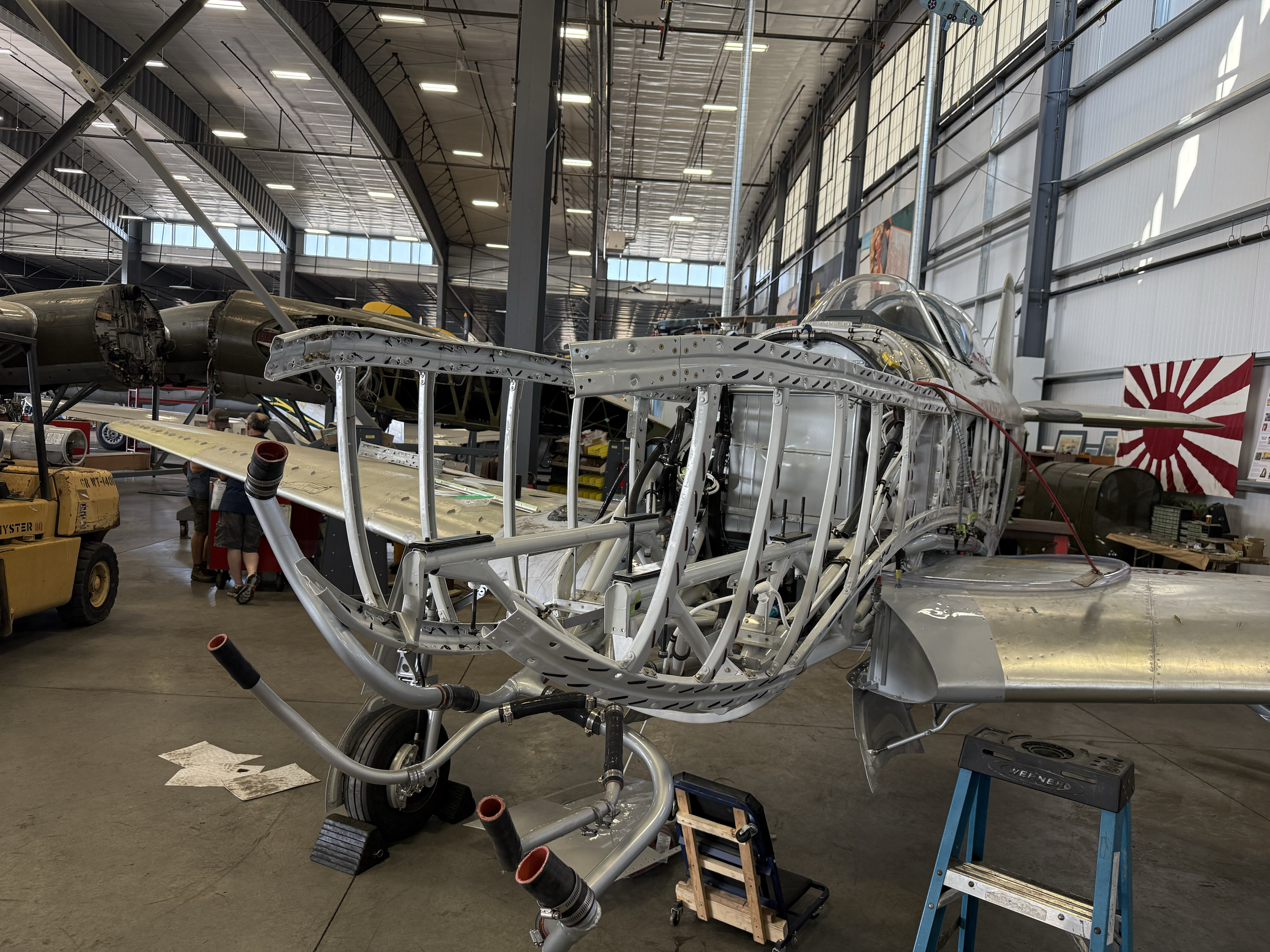 Inside an aircraft manufacturing or restoration hangar, a partially assembled airplane with exposed metal framework and components is visible. The building has high ceilings with skylights and industrial lighting. Several tools and equipment are scat