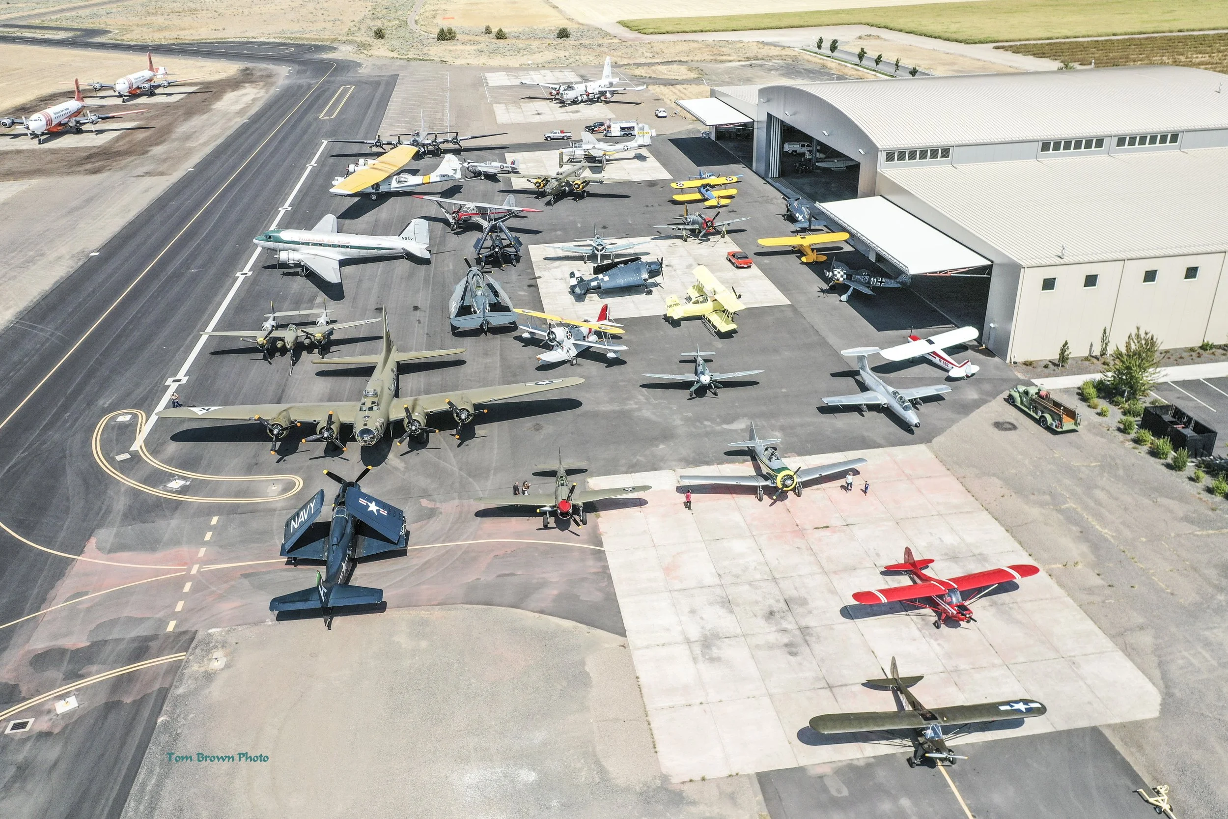 Aerial view of an aircraft storage hangar with various vintage and military planes parked outside, some inside the hangar, with open doors, on a large concrete tarmac.