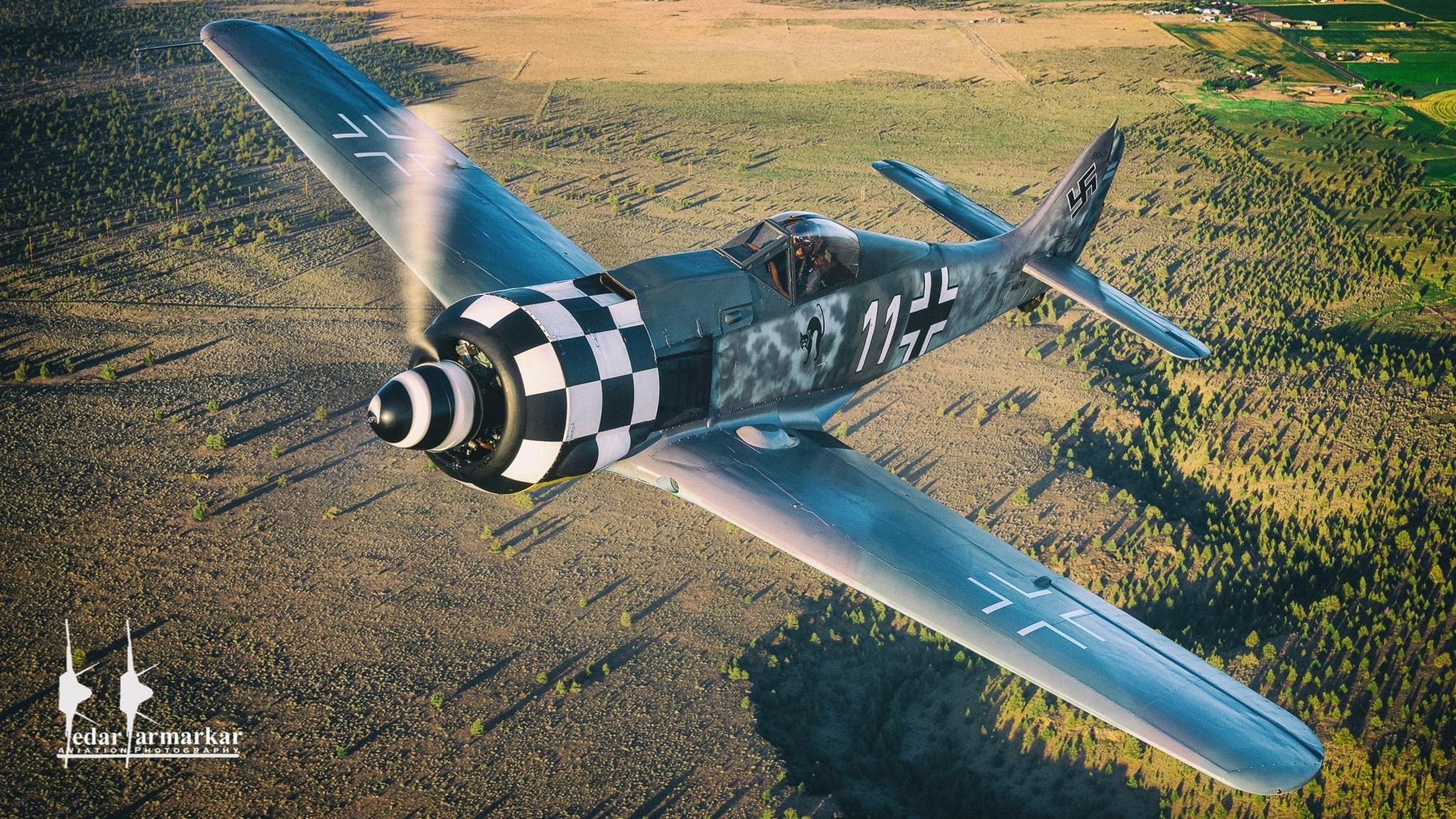 A vintage military fighter plane flying above farmland with fields and scattered trees.