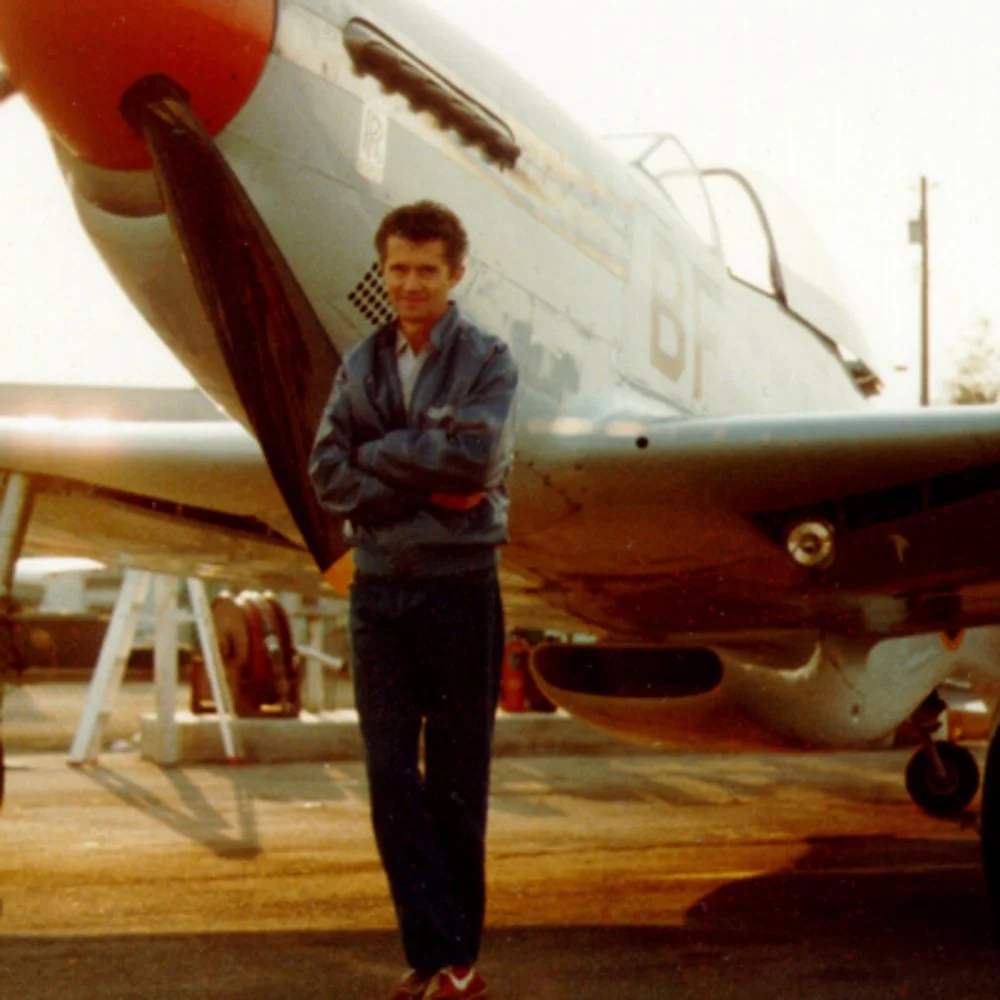 Jack Erickson standing in front of a vintage silver fighter jet aircraft at an airfield, with arms crossed and smiling.