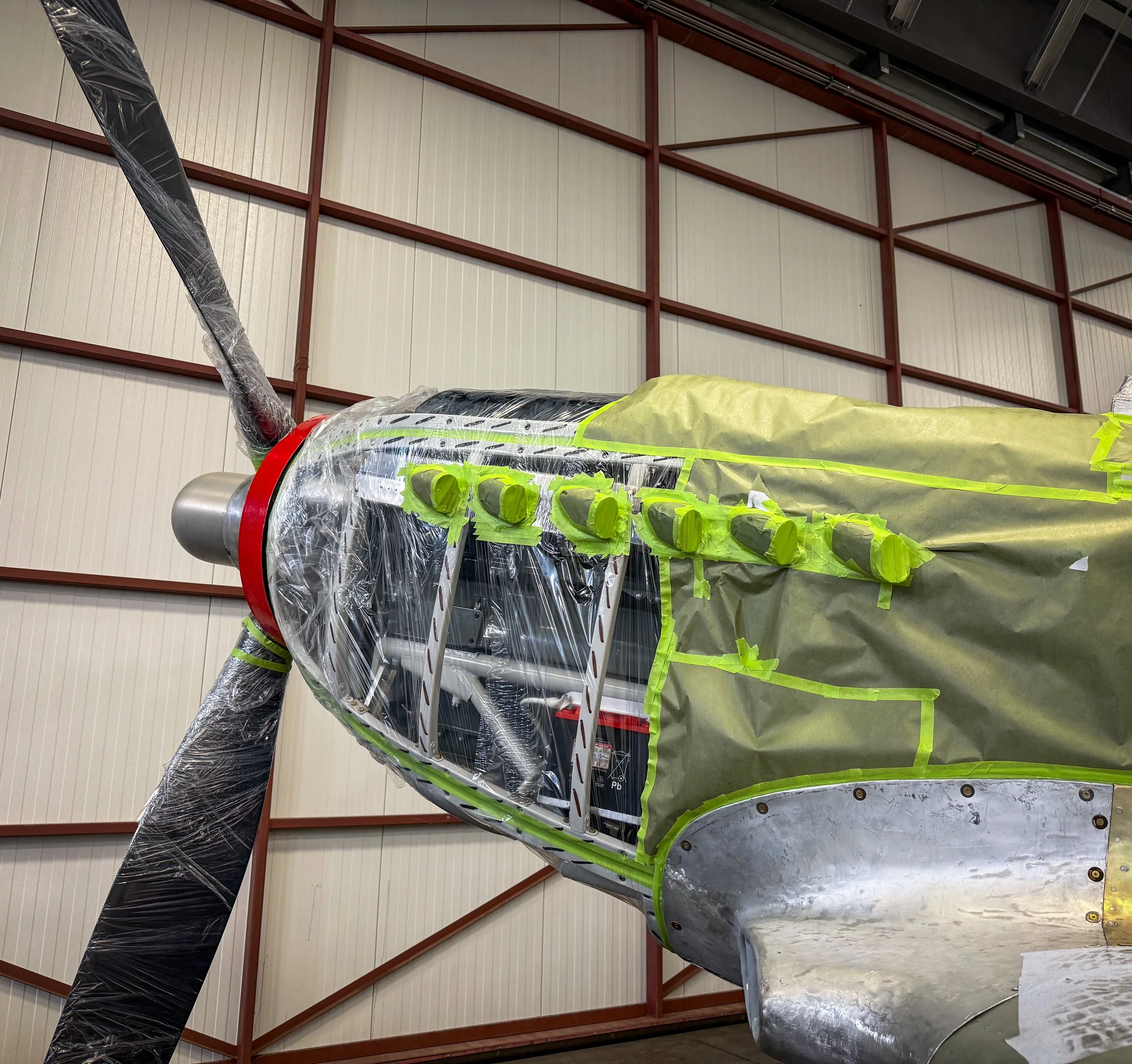 A rocket model wrapped in protective plastic with green tape on parts of it, situated in a large indoor space with metal walls and a ceiling.
