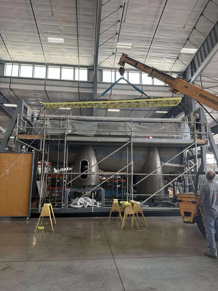 Construction workers and equipment installing or assembling a large stainless steel industrial or scientific vessel inside a spacious warehouse or factory with high ceilings and windows.