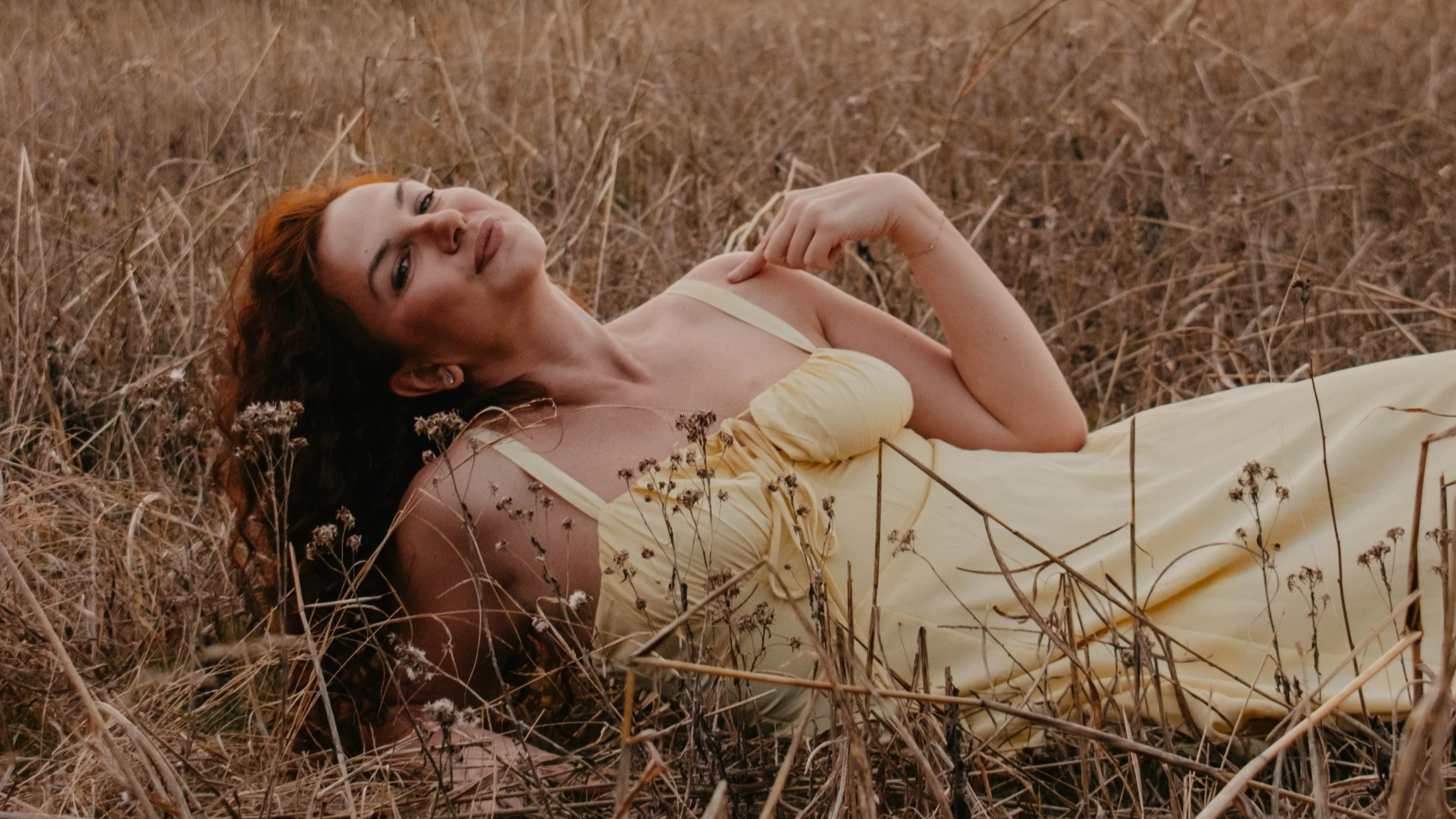 A woman with brown curly hair lying in a field of dry grass, wearing a light yellow dress with spaghetti straps.