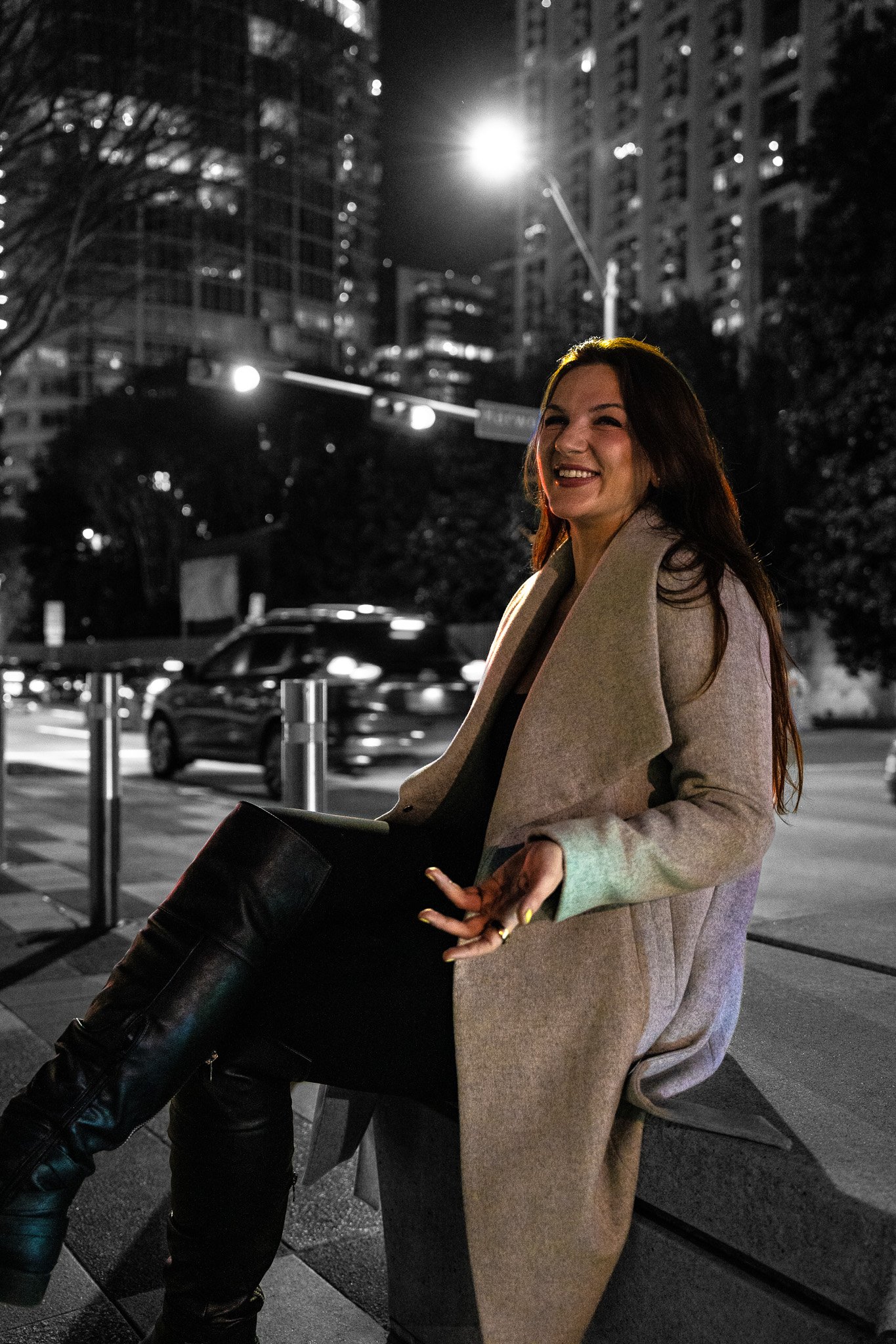 A woman sitting on a bench at night in an urban setting with tall buildings, cars, and streetlights around her, smiling and enjoying the evening.