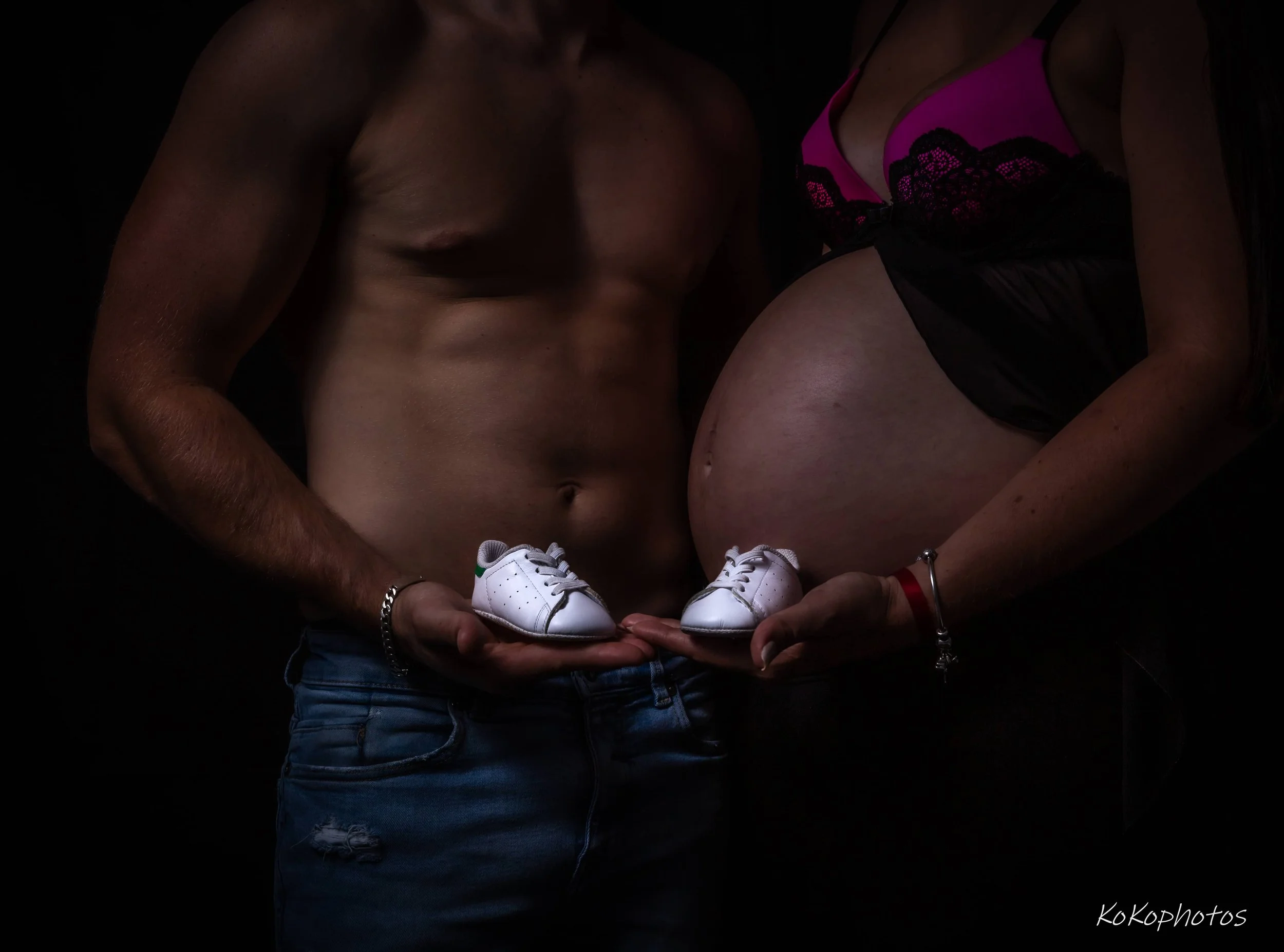 Two women are holding a pair of small white shoes in front of their bodies, with one woman shirtless and the other wearing pink and black lingerie, against a dark background.