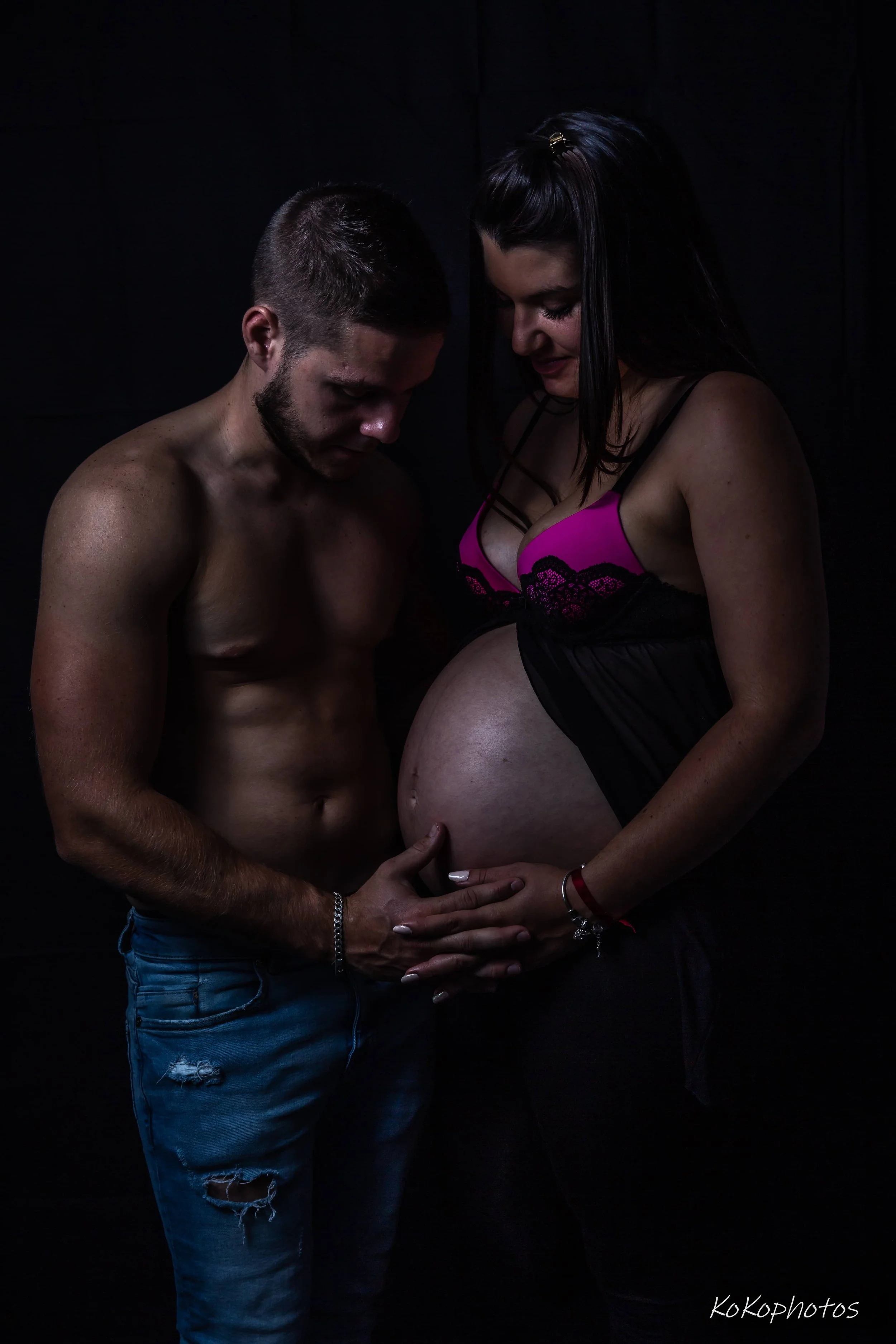 A shirtless man with facial hair and ripped jeans holding a pregnant woman’s pregnant belly, both with their heads bowed and eyes closed, touching her belly gently in a dark setting.