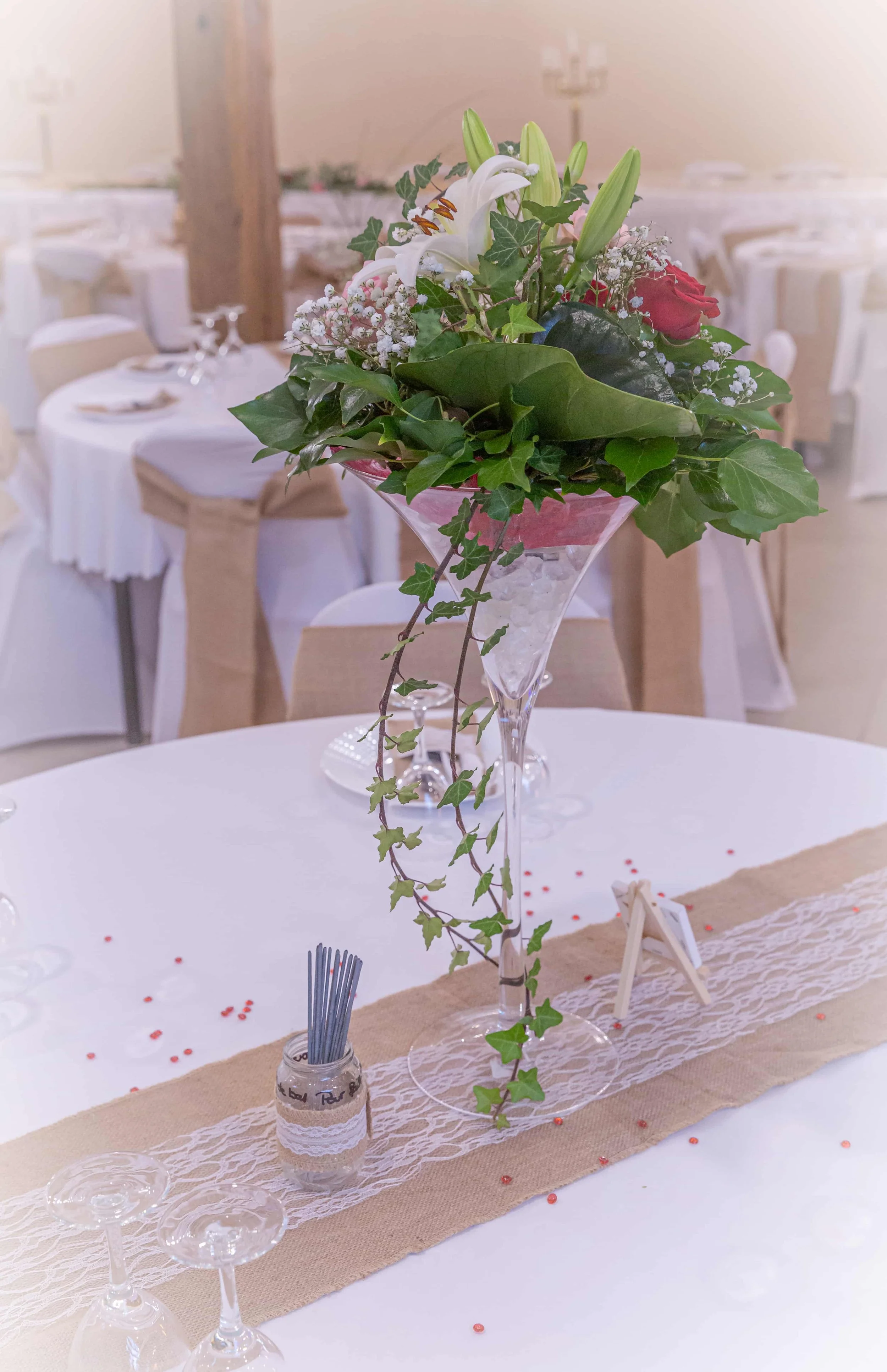 A tall glass vase with a floral arrangement of white lilies, a pink rose, and green ivy on a banquet table decorated with lace and burlap runner, surrounded by white chairs in a decorated banquet hall.