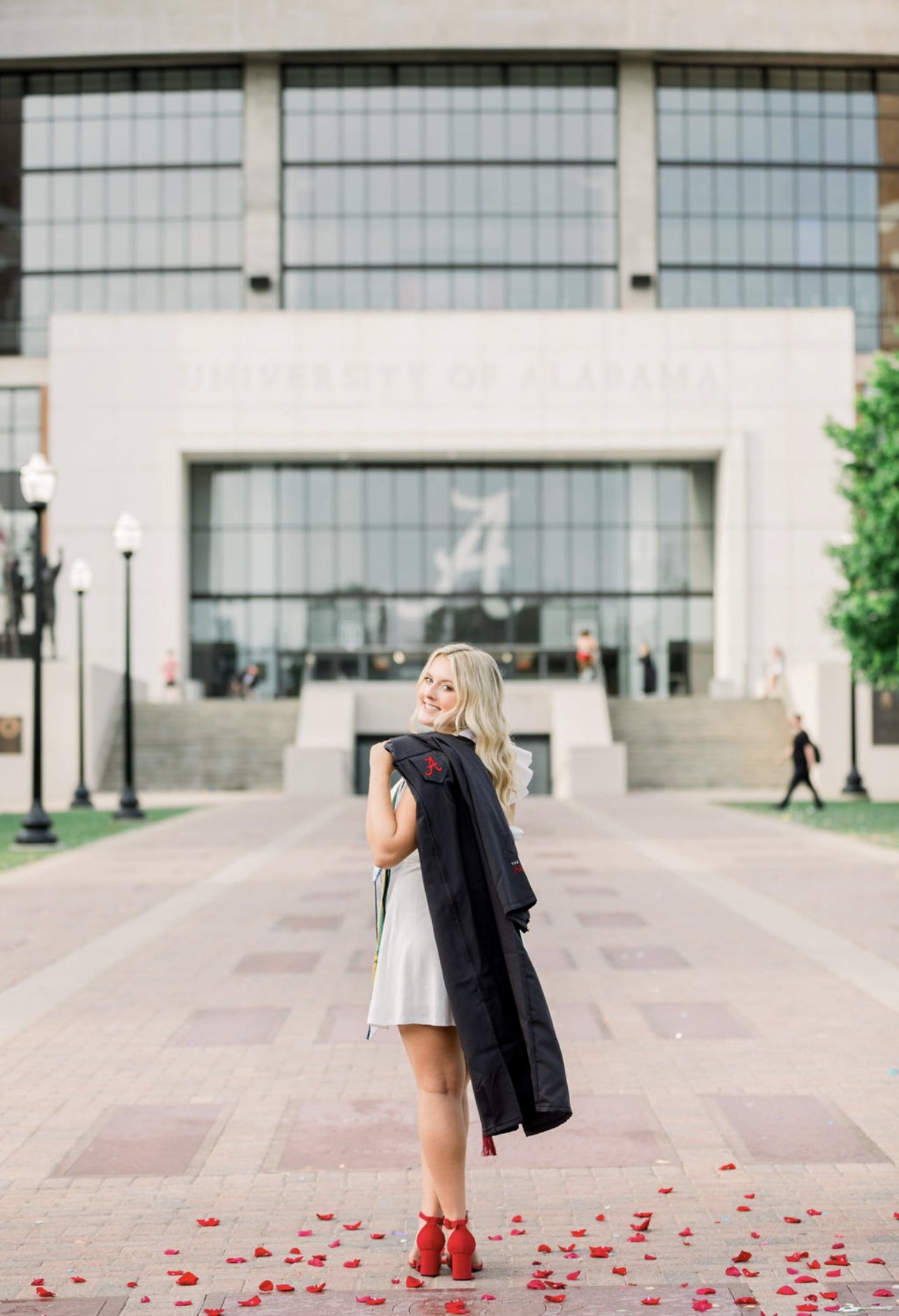 A young woman with blonde hair in a white dress and red high heels walks on a sidewalk covered with rose petals, carrying a black graduation gown on her shoulder, in front of a university building.