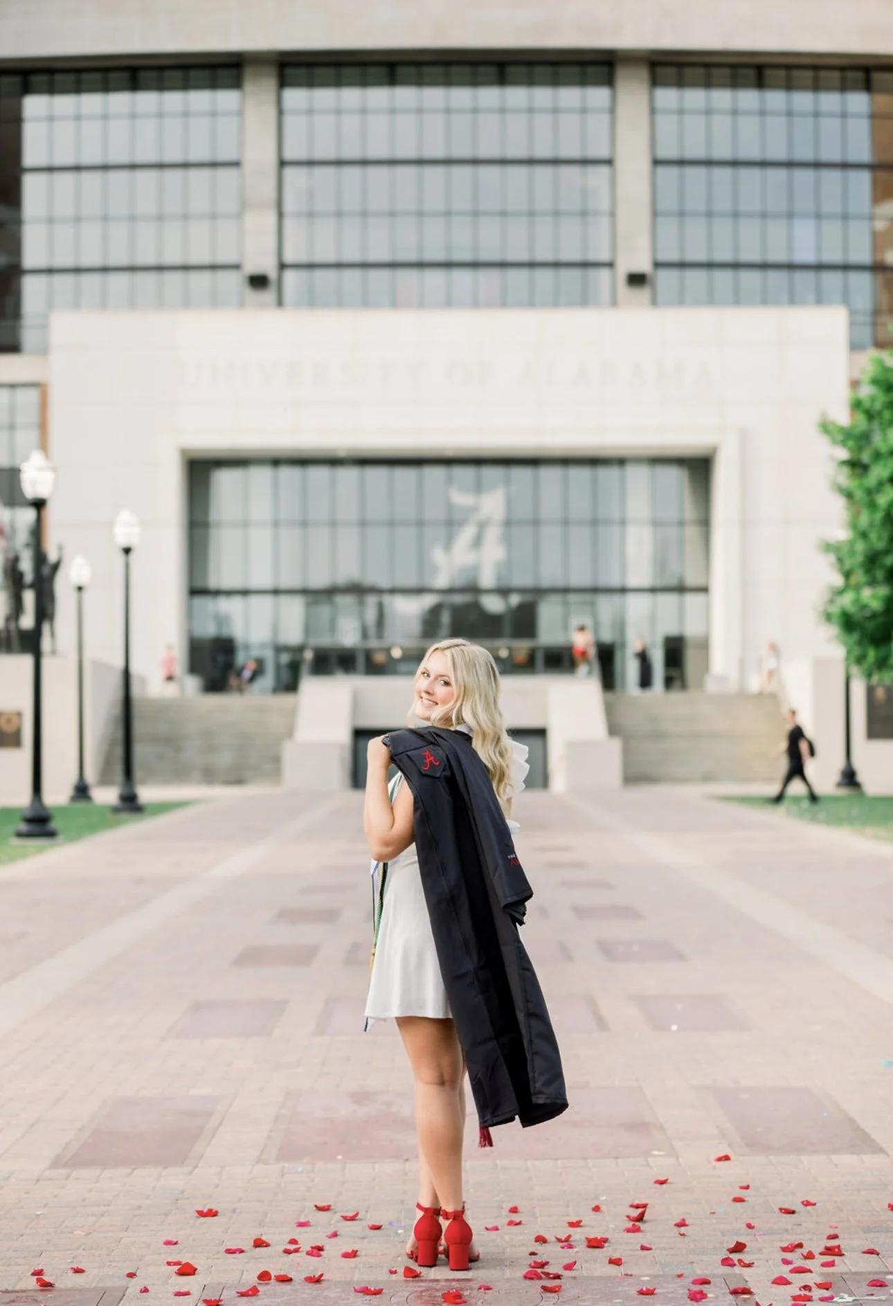 A young woman in a white dress and red heels stands on a paved walkway outside a university building, holding a graduation gown over her shoulder and smiling at the camera. Red flower petals are scattered on the ground around her.