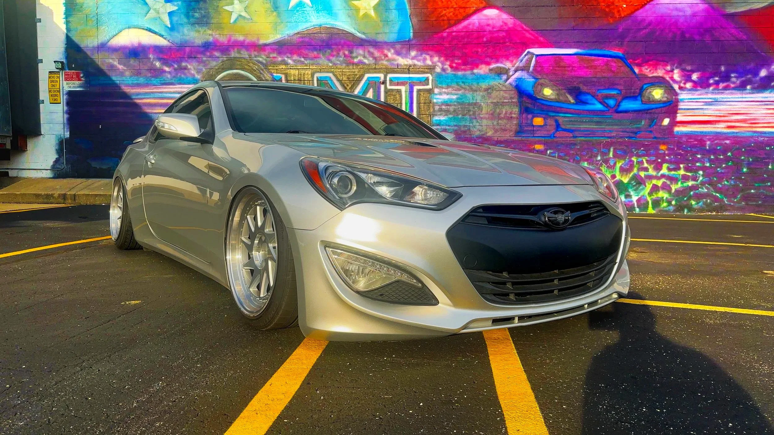 A silver car parked in a parking lot with a colorful mural of a sports car, mountains, and stars in the background.