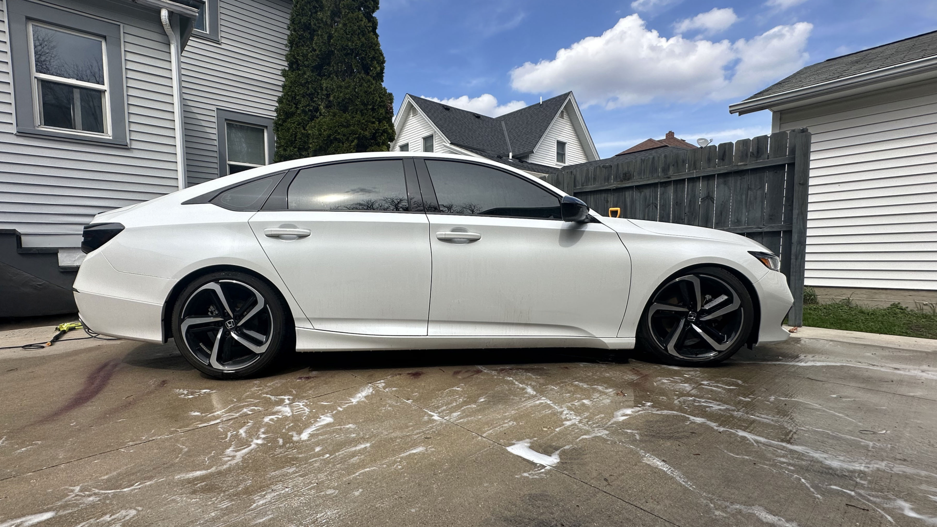 White sedan car parked on a concrete driveway near a wooden fence, with houses and a cloudy sky in the background.