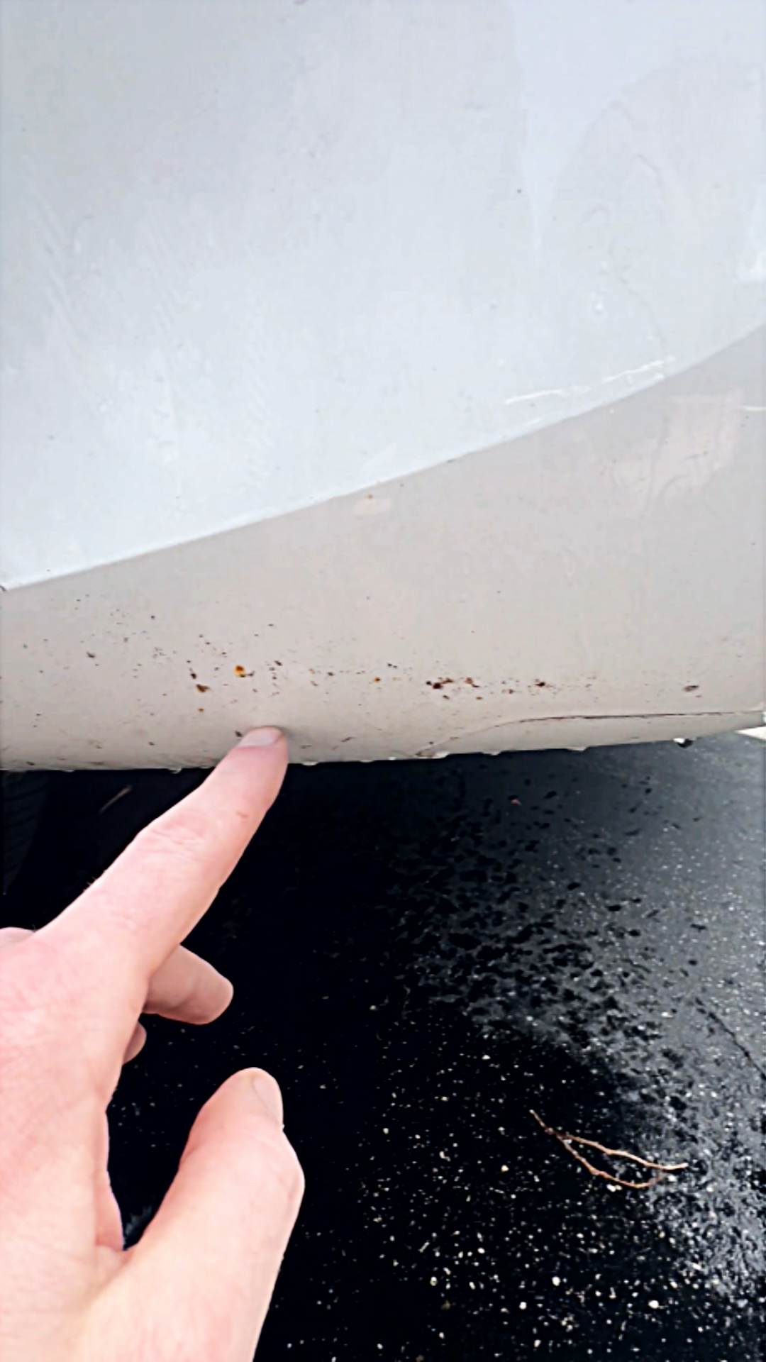 Close-up of a person's finger pointing at dirt and grime on the lower part of a white vehicle's bumper, with a black asphalt ground below.