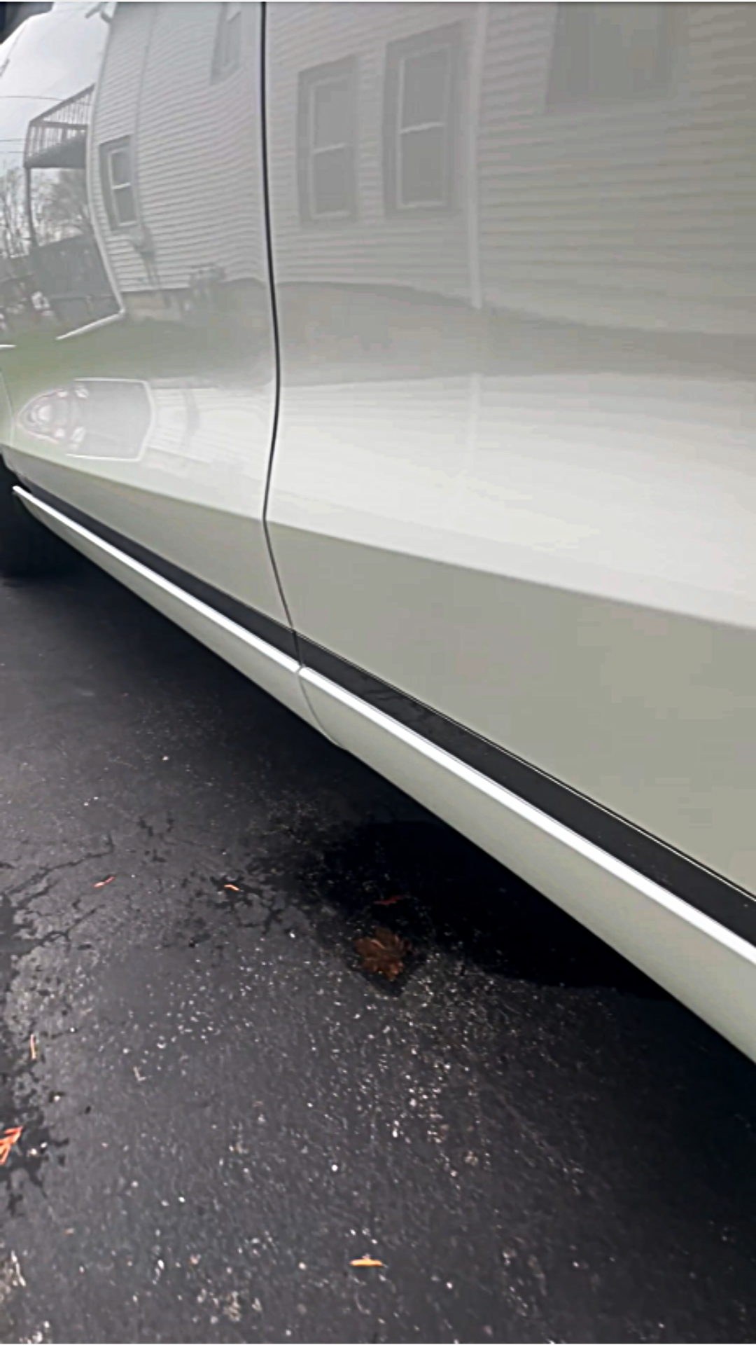 Close-up of a beige car's side panel and door, reflecting houses and trees in the background on a paved asphalt surface.