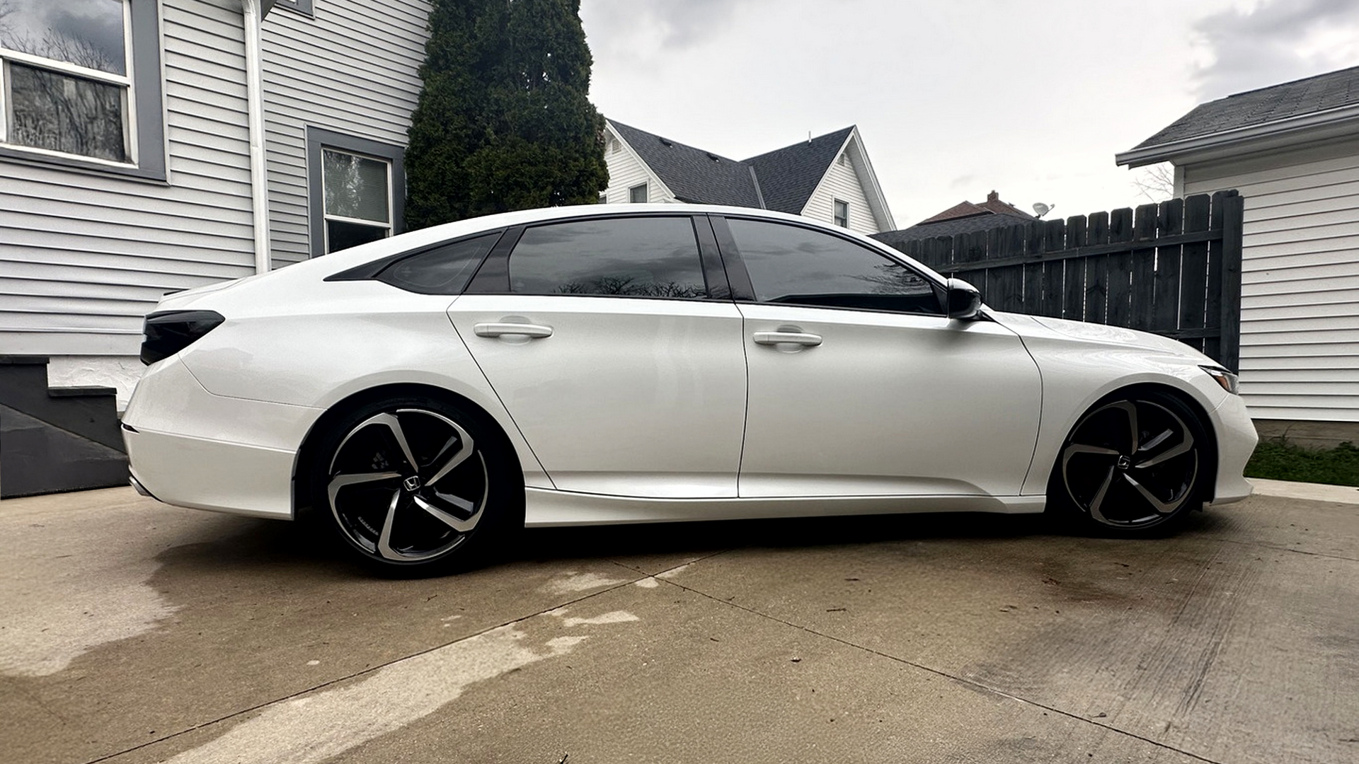 Side view of a white modern sedan car parked on a driveway in front of residential houses with vinyl siding, a black fence, and overcast sky.