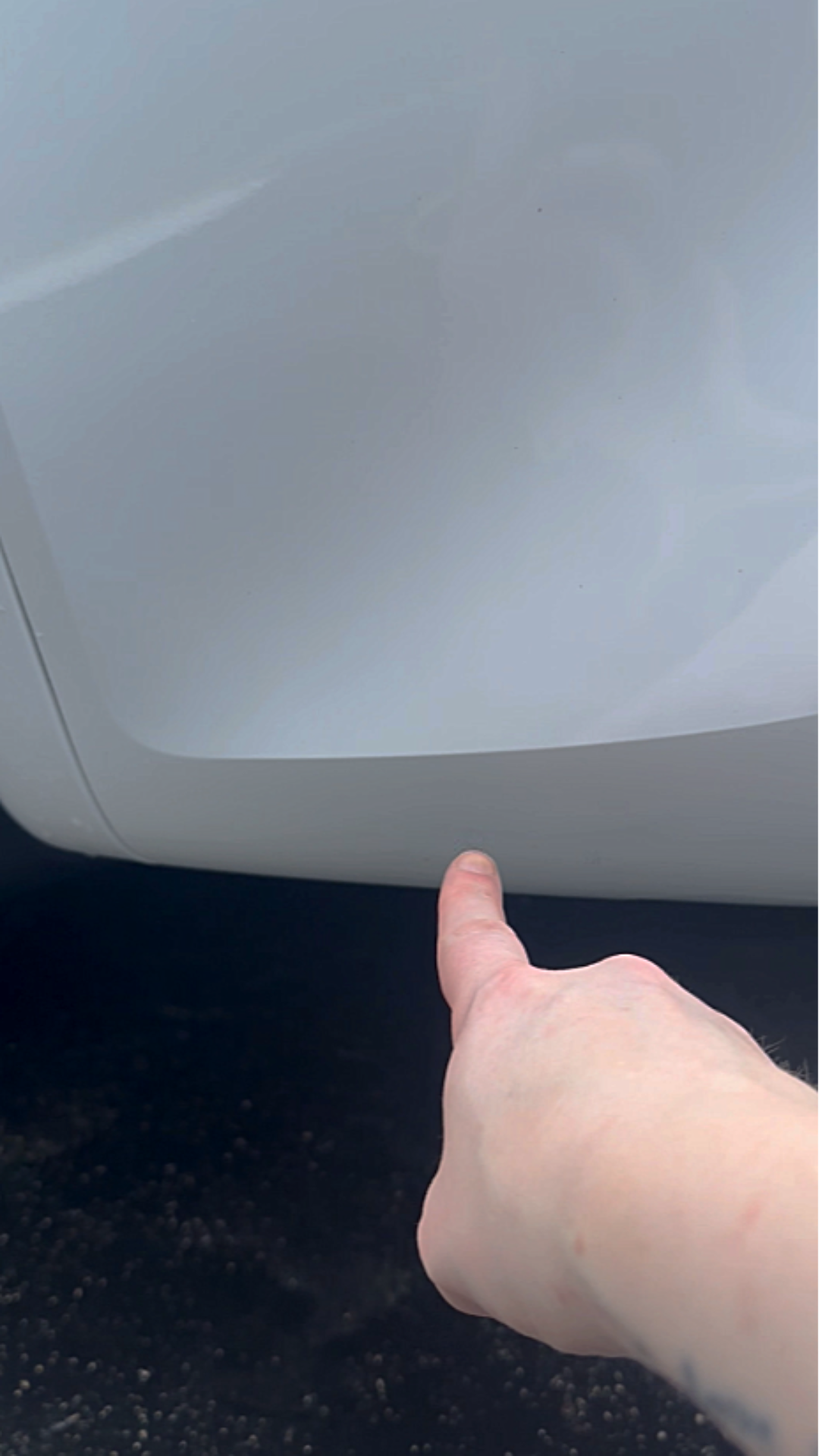 A person pointing at the corner of a white appliance, possibly a washing machine or dryer, with a dark textured floor beneath.