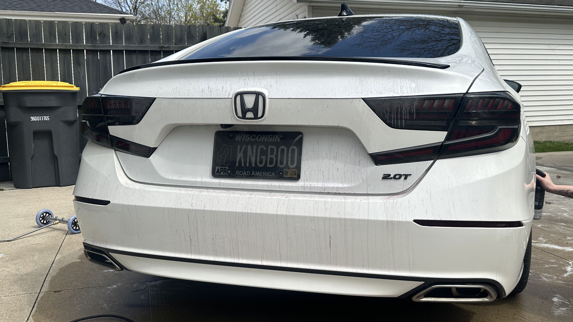 White Honda sedan with Wisconsin license plate and black taillights, parked in a driveway with a washing tool on the side.