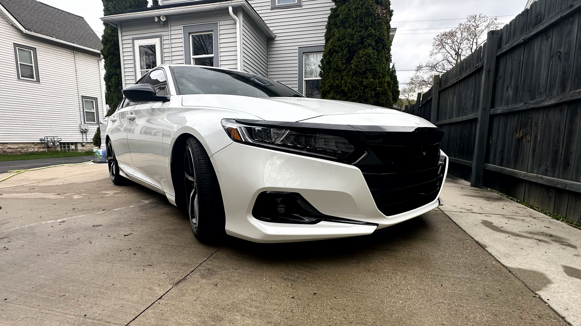 White sedan car parked in driveway with black grille and black accents, house and wooden fence in background.