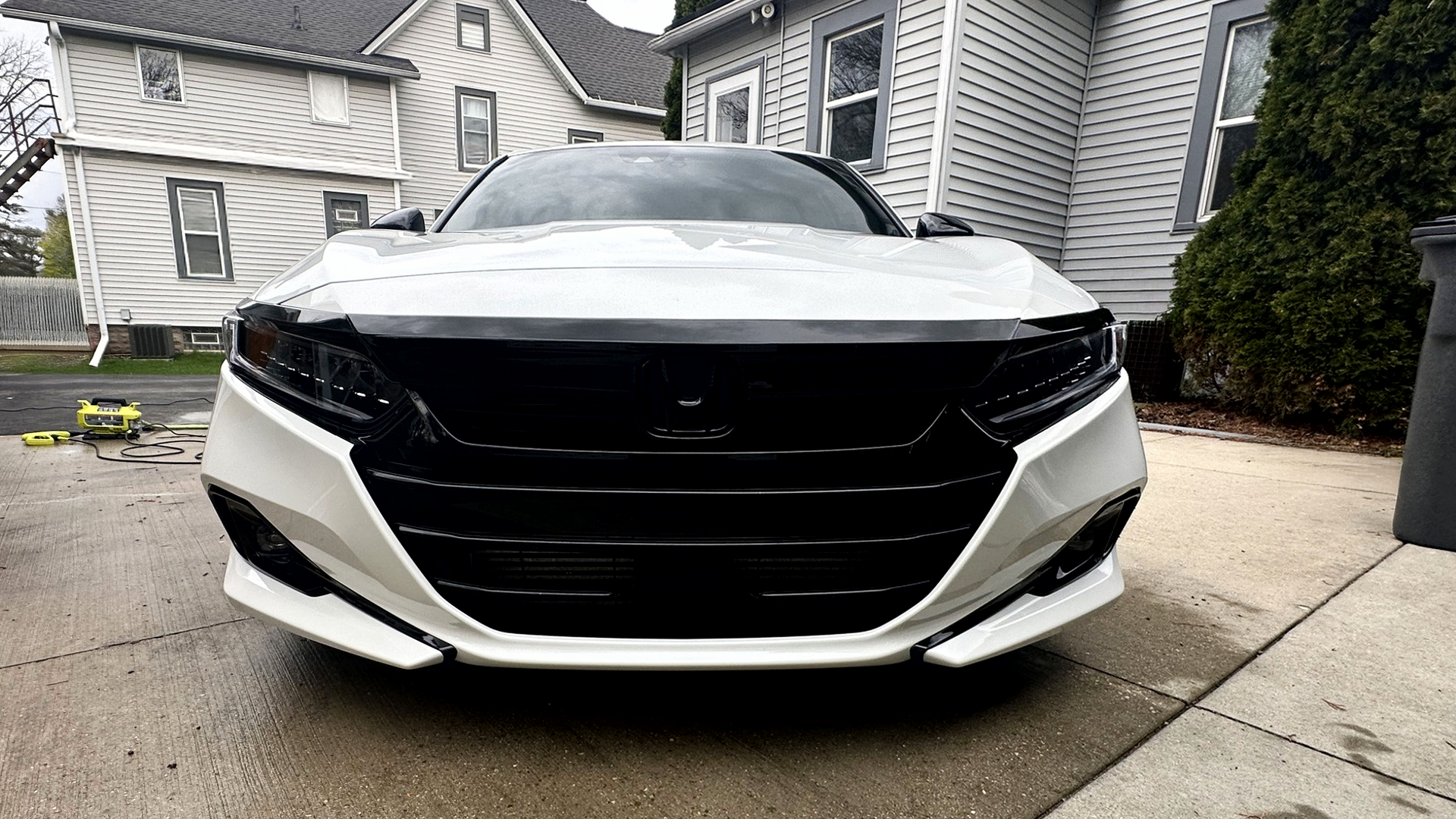 Front view of a white car with black grille and headlights parked on a concrete driveway in a residential area, with houses and greenery in the background.