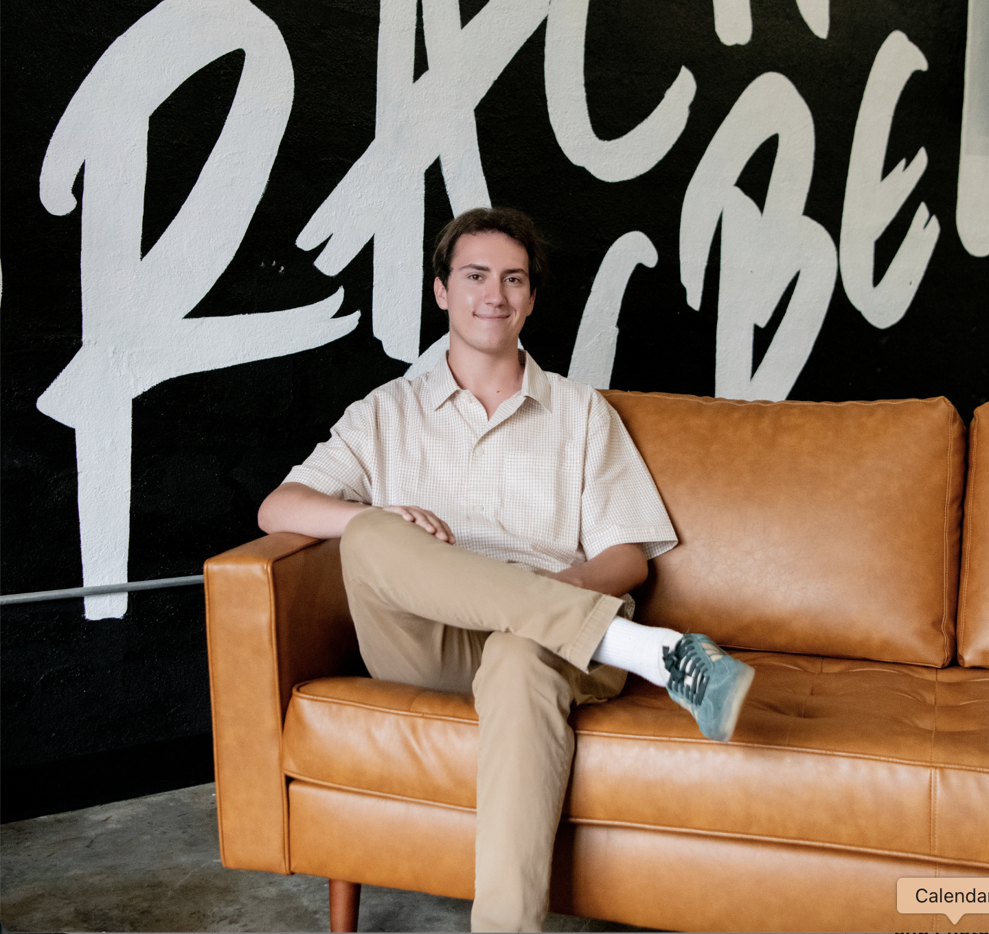 A young man with dark hair, wearing a beige checkered shirt, beige pants, and teal sneakers, sitting cross-legged on a tan leather couch in front of a black wall with large white handwritten-style lettering.