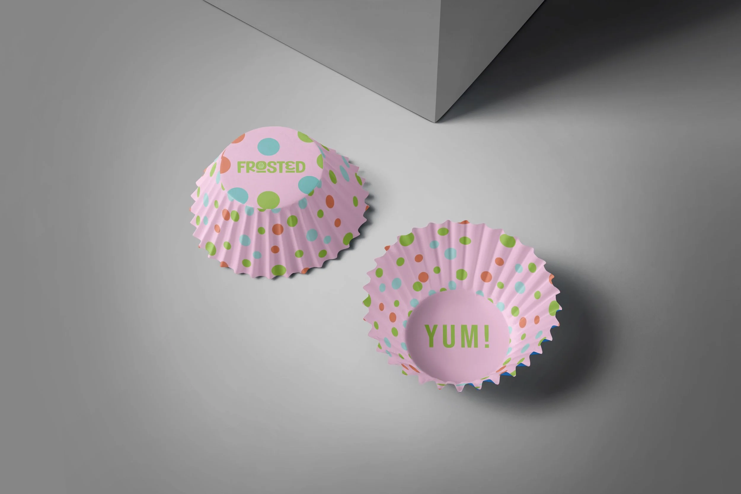 Colorful cupcake liners with polka dot patterns, with the words 'Frosted' and 'Yum!' printed on two of the liners, resting on a gray surface with a corner of a gray box visible in the background.