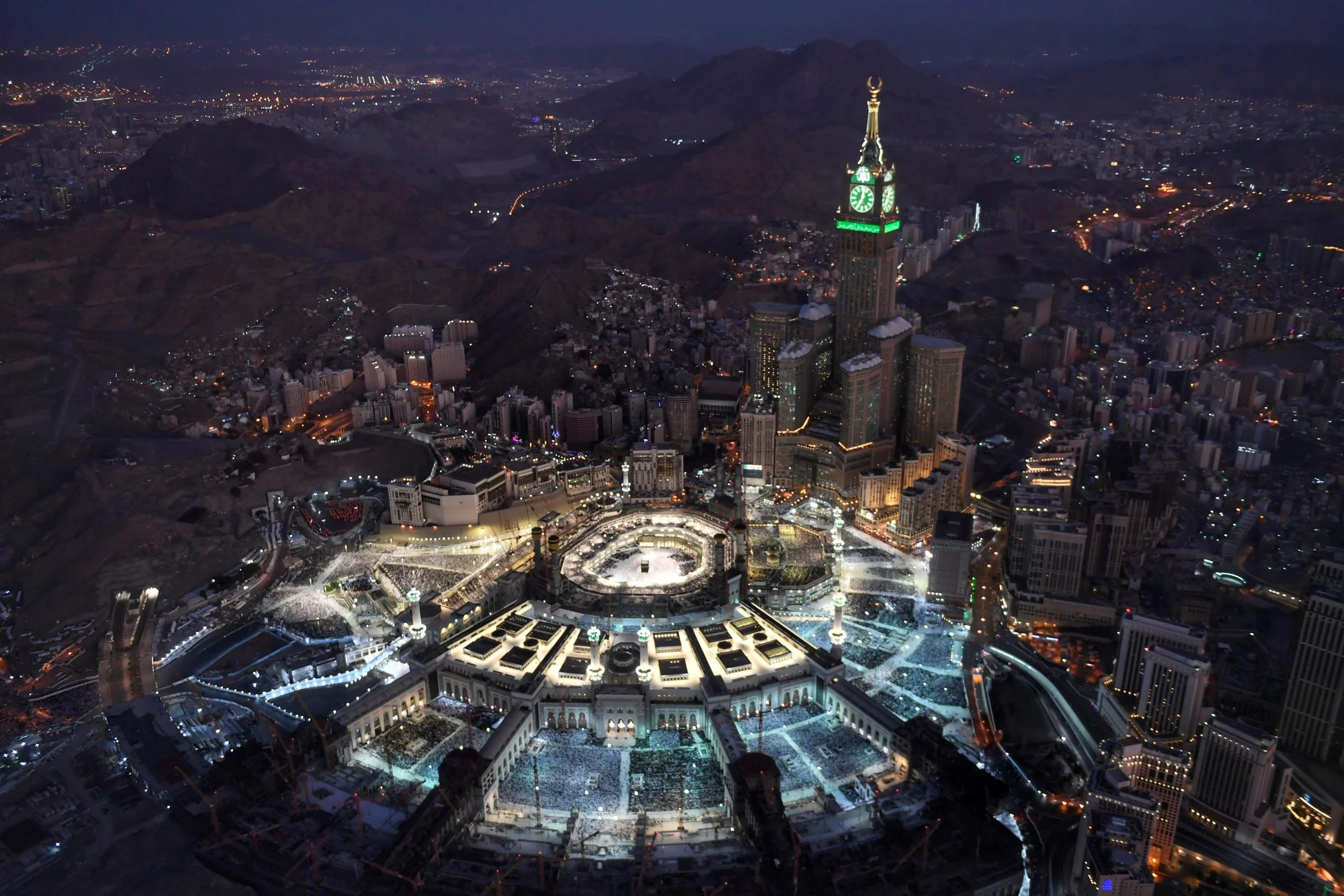 Aerial night view of Masjid al-Haram in Makkah with the Kaaba at the center, surrounded by city lights, mountain ranges, and the Abraj Al Bait Clock Tower.