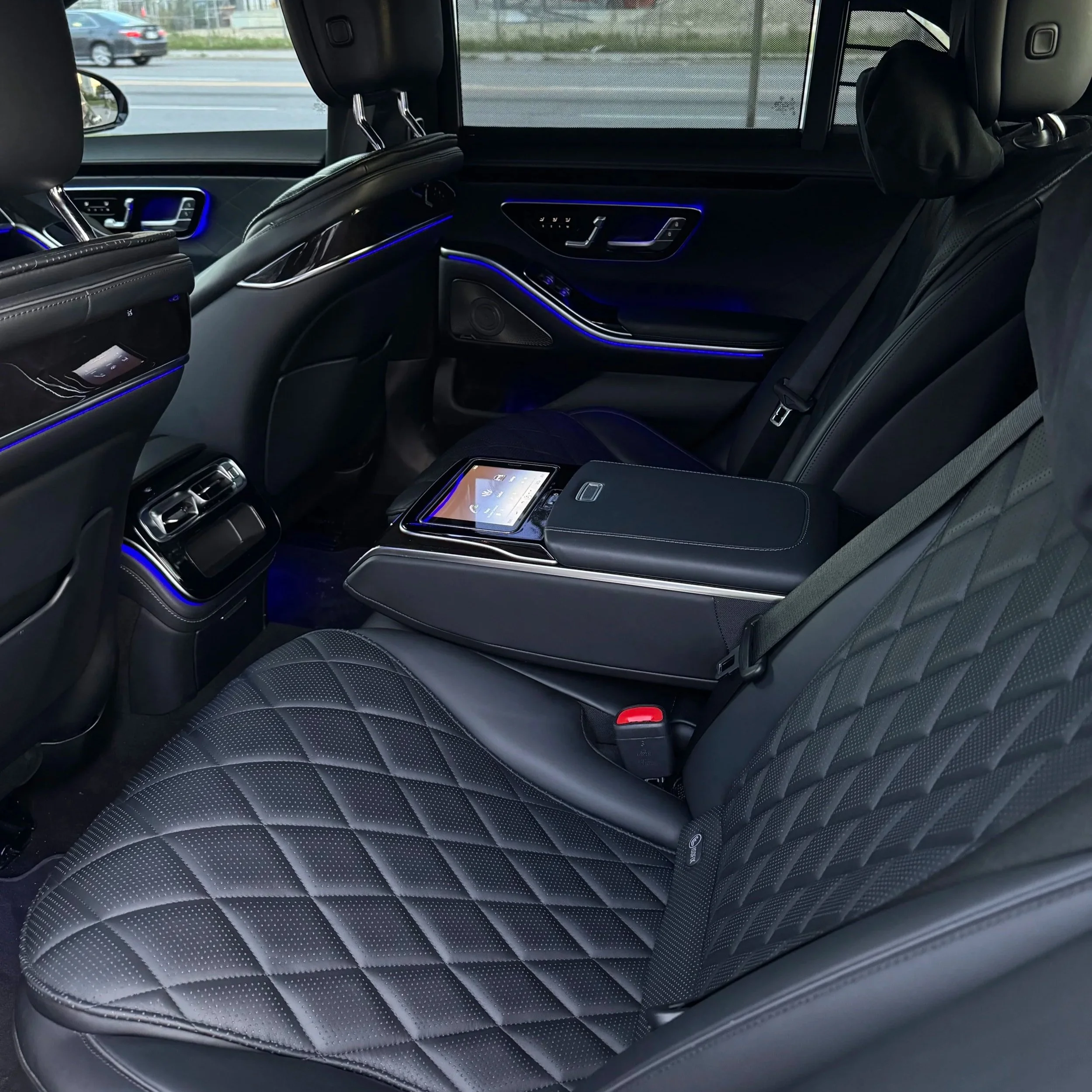 Interior of a luxury car with black leather seats with diamond stitching, illuminated ambient lighting, and a touchscreen control panel on the armrest of the rear seat.