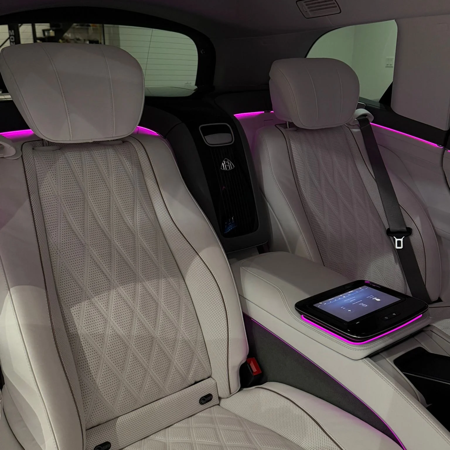 The interior of a luxury car with beige leather seats, illuminated pink accent lighting along the headrests and side panels, a touchscreen control panel on the center console, and seat belts.