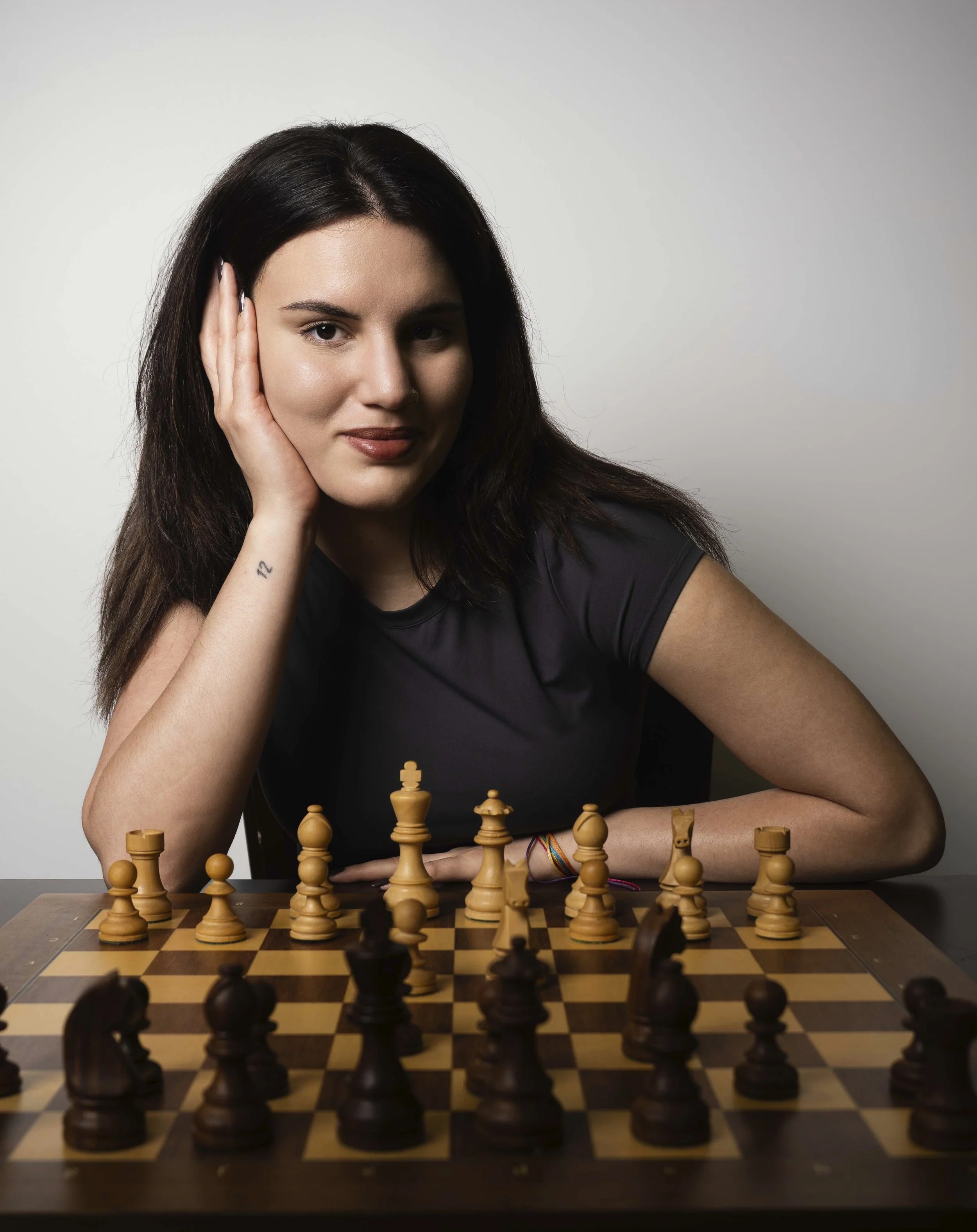 Young woman with dark hair and a nose piercing playing chess, seated at a table with a chessboard and wooden chess pieces, against a plain gray background.