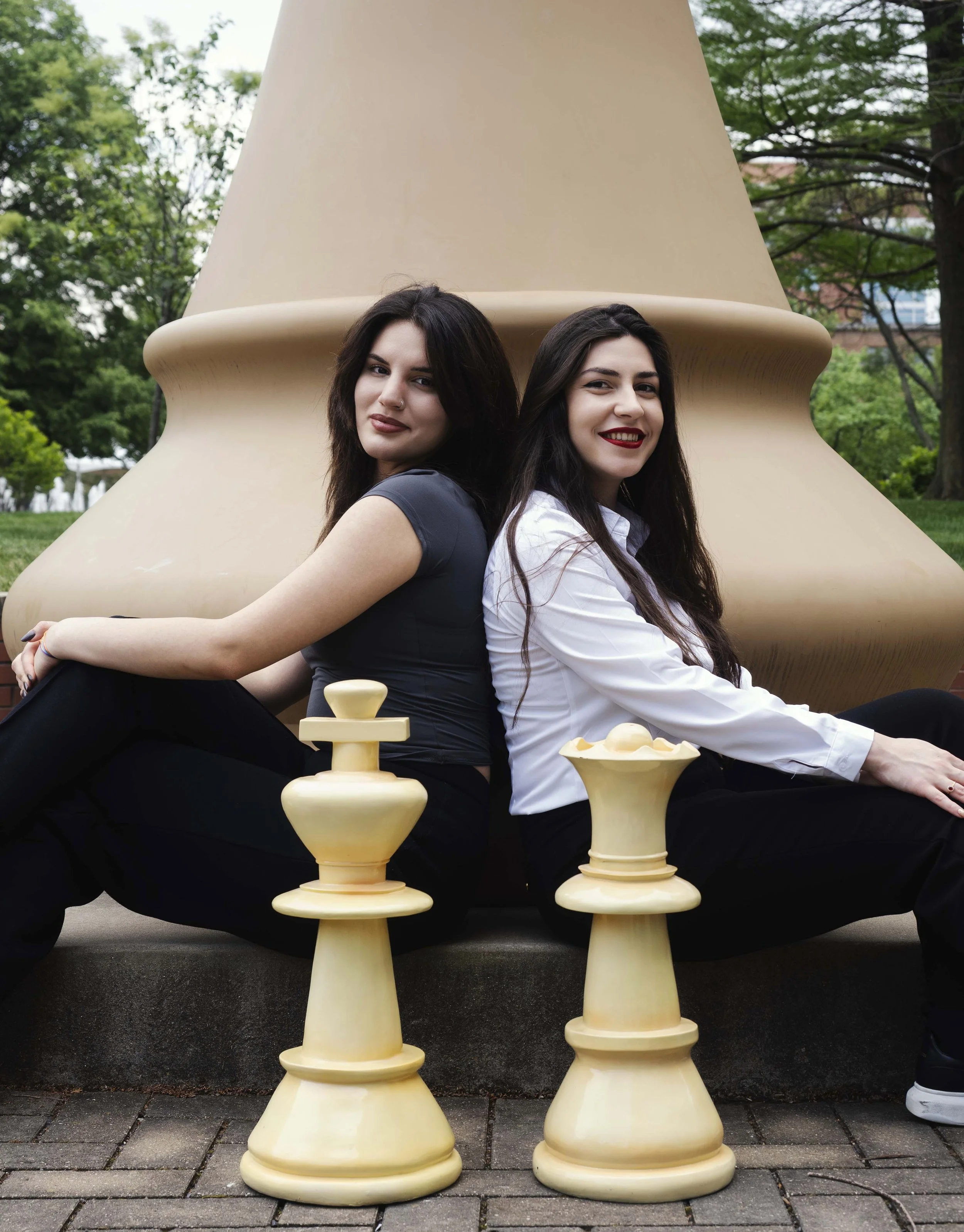 Two women sitting back-to-back on large beige chess pieces outdoors, with green trees and buildings in the background.